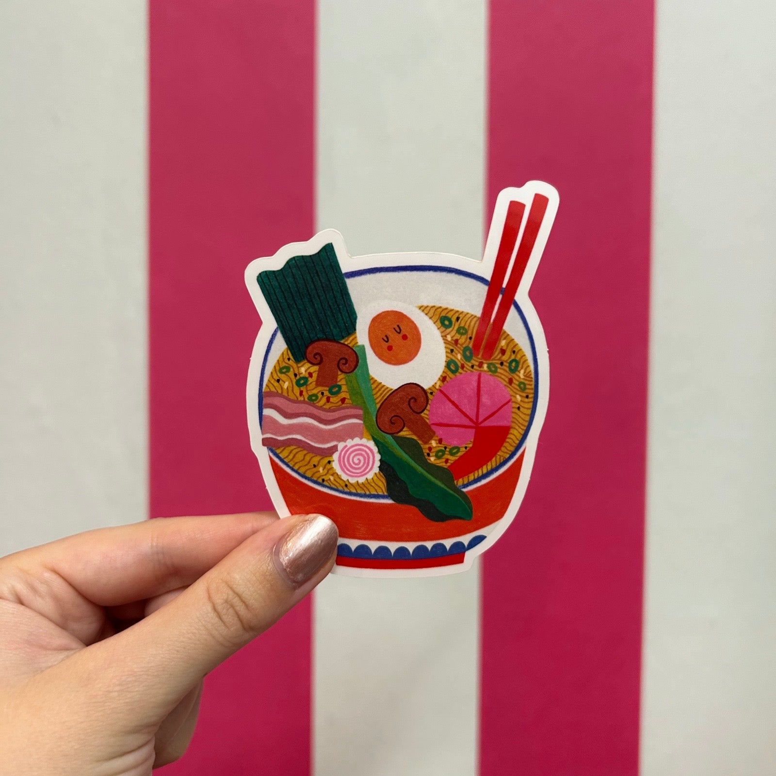 A hand with metallic pink nail polish holds a sticker of a colourful ramen bowl against a background of vertical pink and white stripes.