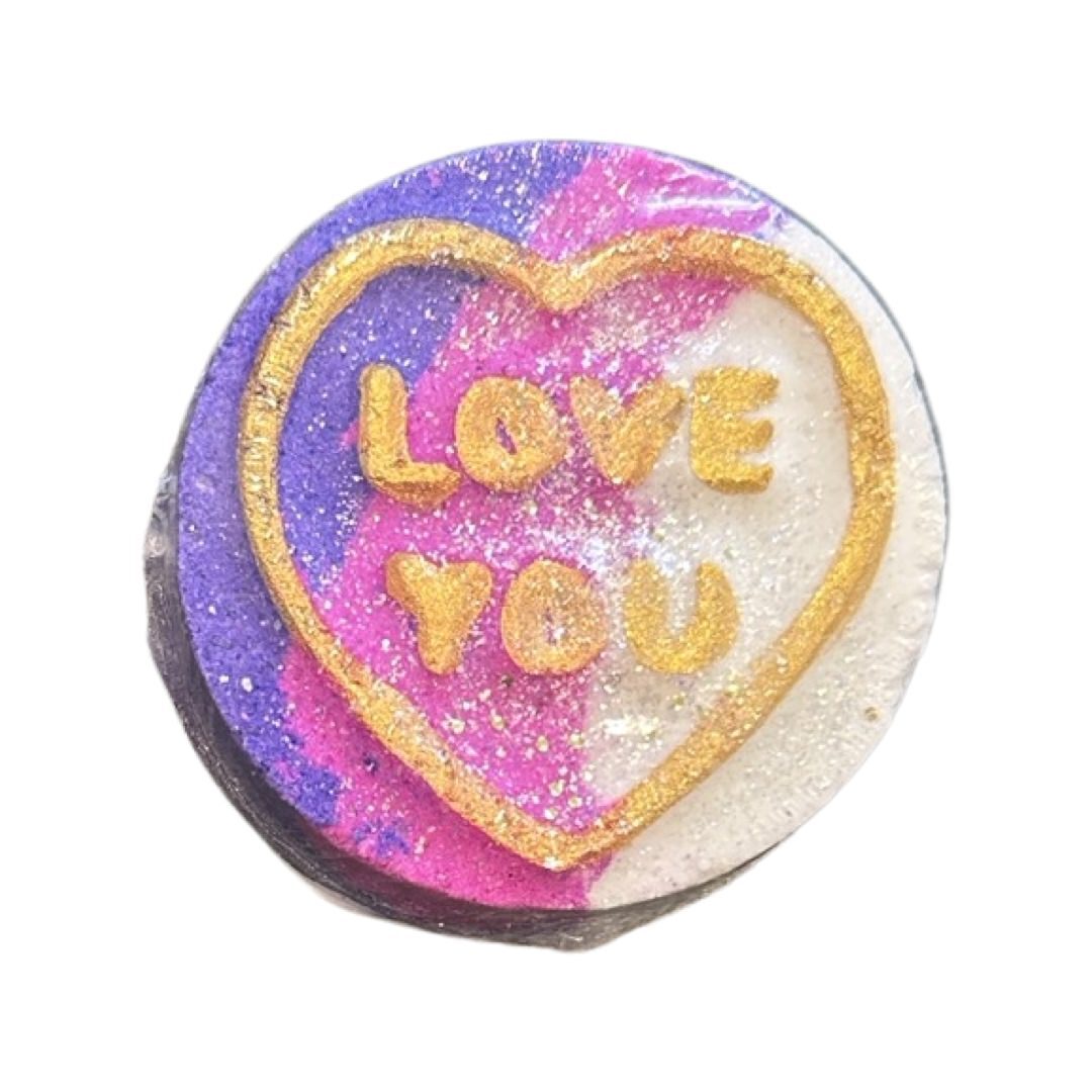 A circular bath bomb inspired by the retro love hearts sweets in pink, purple and white with the text 'Love You' written in gold.