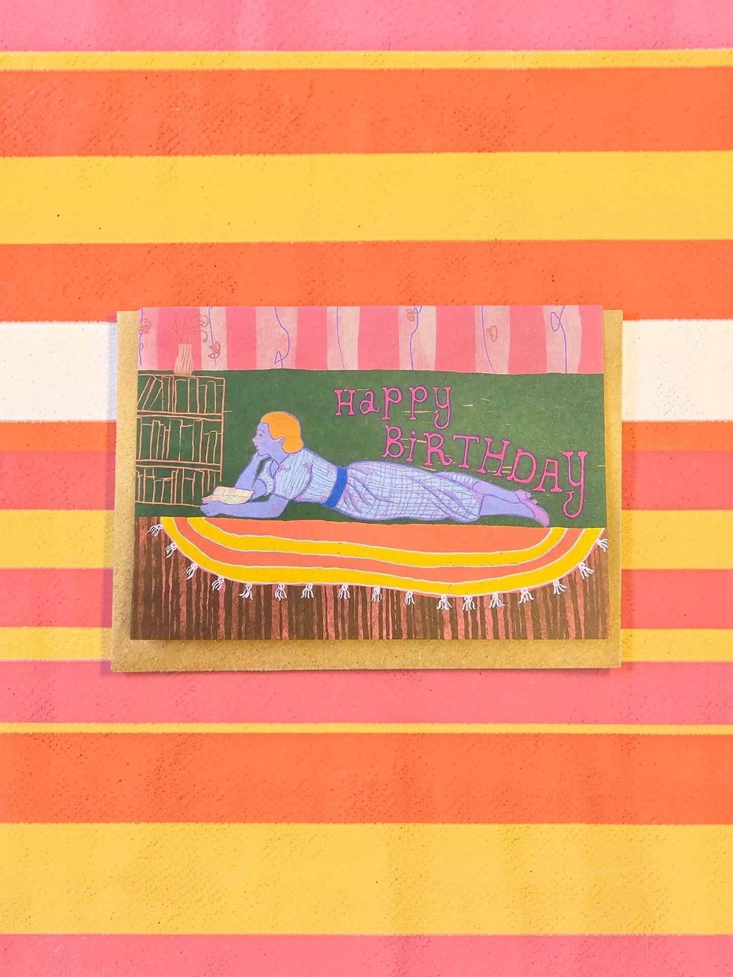 Birthday card with a person lying on a rug and 'Happy Birthday' text, on a striped background.