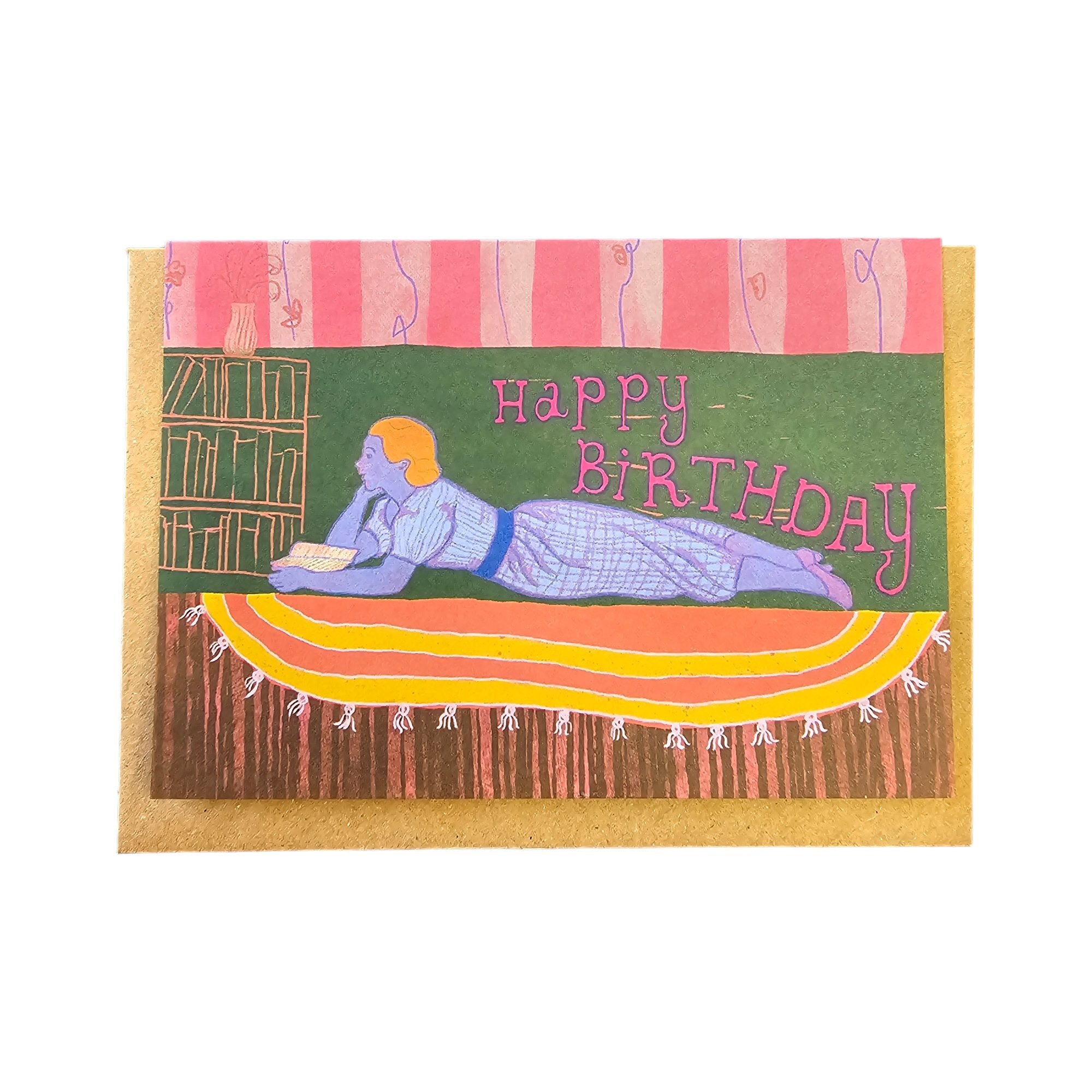 Birthday card featuring a lady with orange hair, dressed in a light blue dress lying on a yellow and orange circular rug reading her favourite book with the text 'Happy Birthday'.