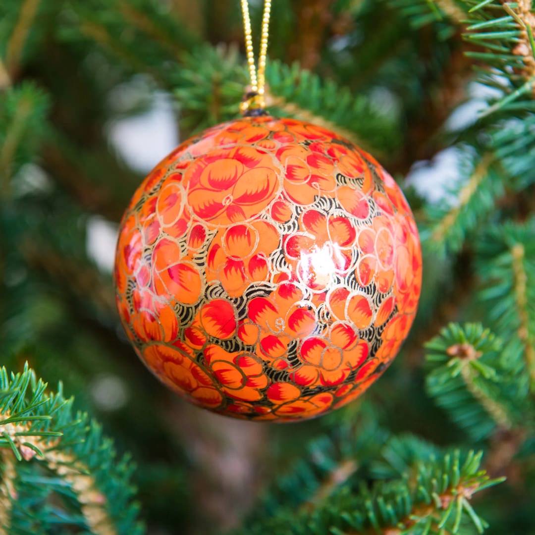 A round papier mache bauble in black with a gold and red floral design hanging on a Christmas tree.