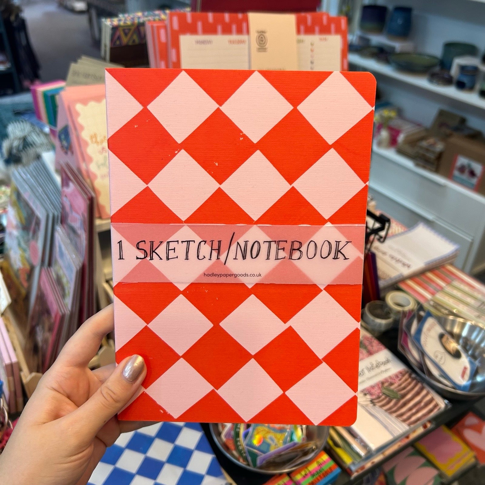 A hand holds a red and pink diamond-patterned sketchbook in a retail store. The cover has a semi-transparent label reading '1 SKETCH/NOTEBOOK'.