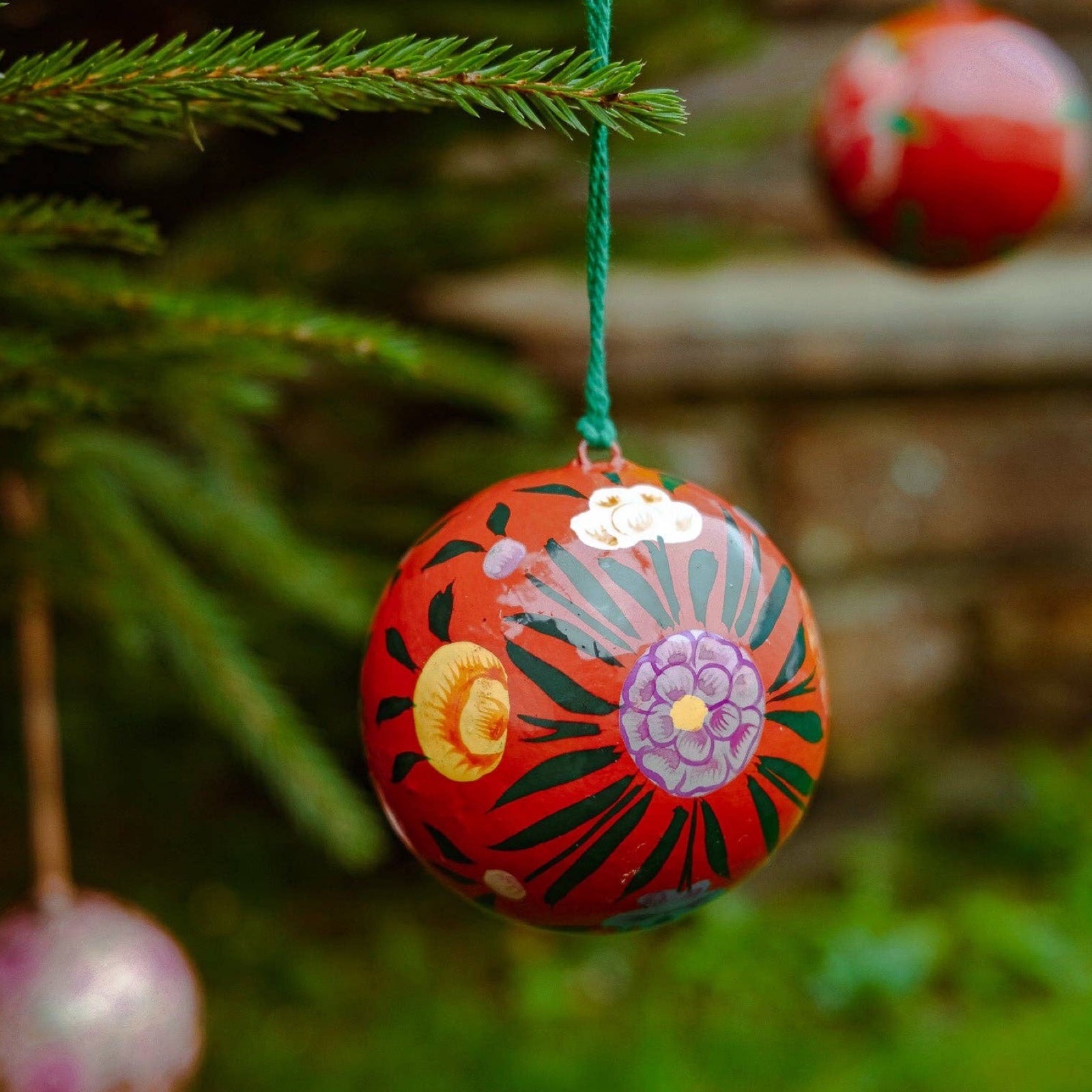 A round papier mache bauble in red with a pink, green, yellow and white floral design hanging on a Christmas tree.