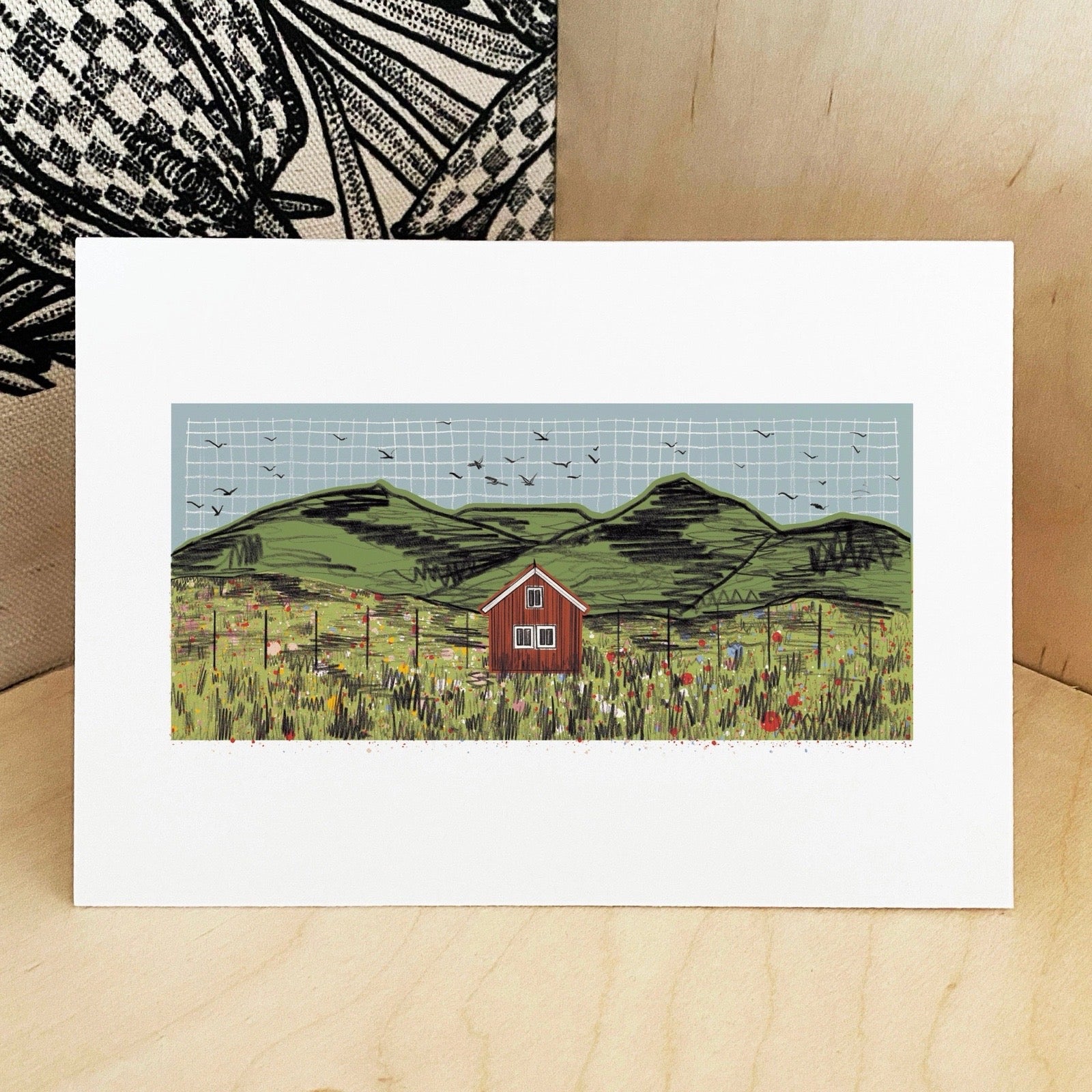 Greeting card with a red cabin in a field design on a wooden surface.
