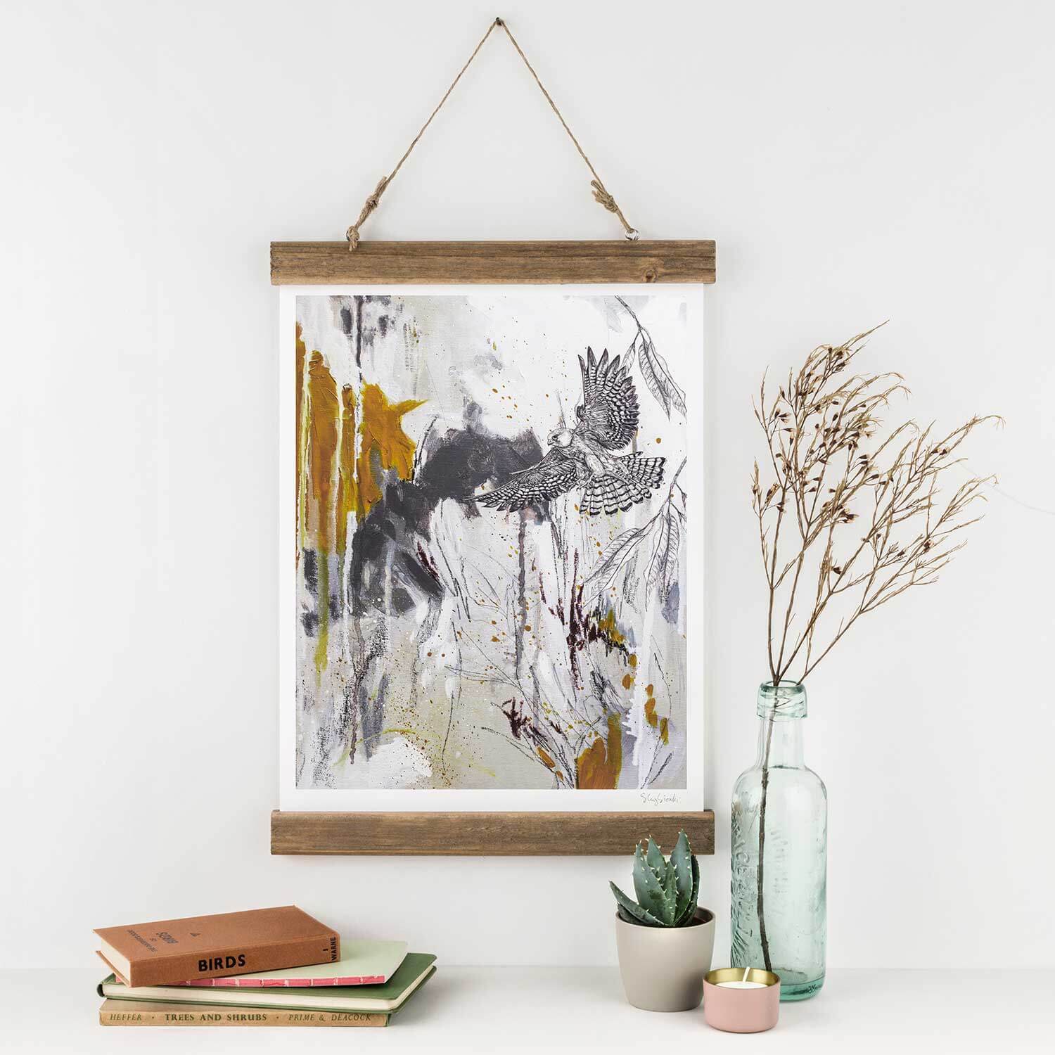 A giclee print of a red footed falcon in flight, with mustard and grey splashes of colour, displayed on a white wall above a potted plant and a glass bottle, with books and a decorative vase below.