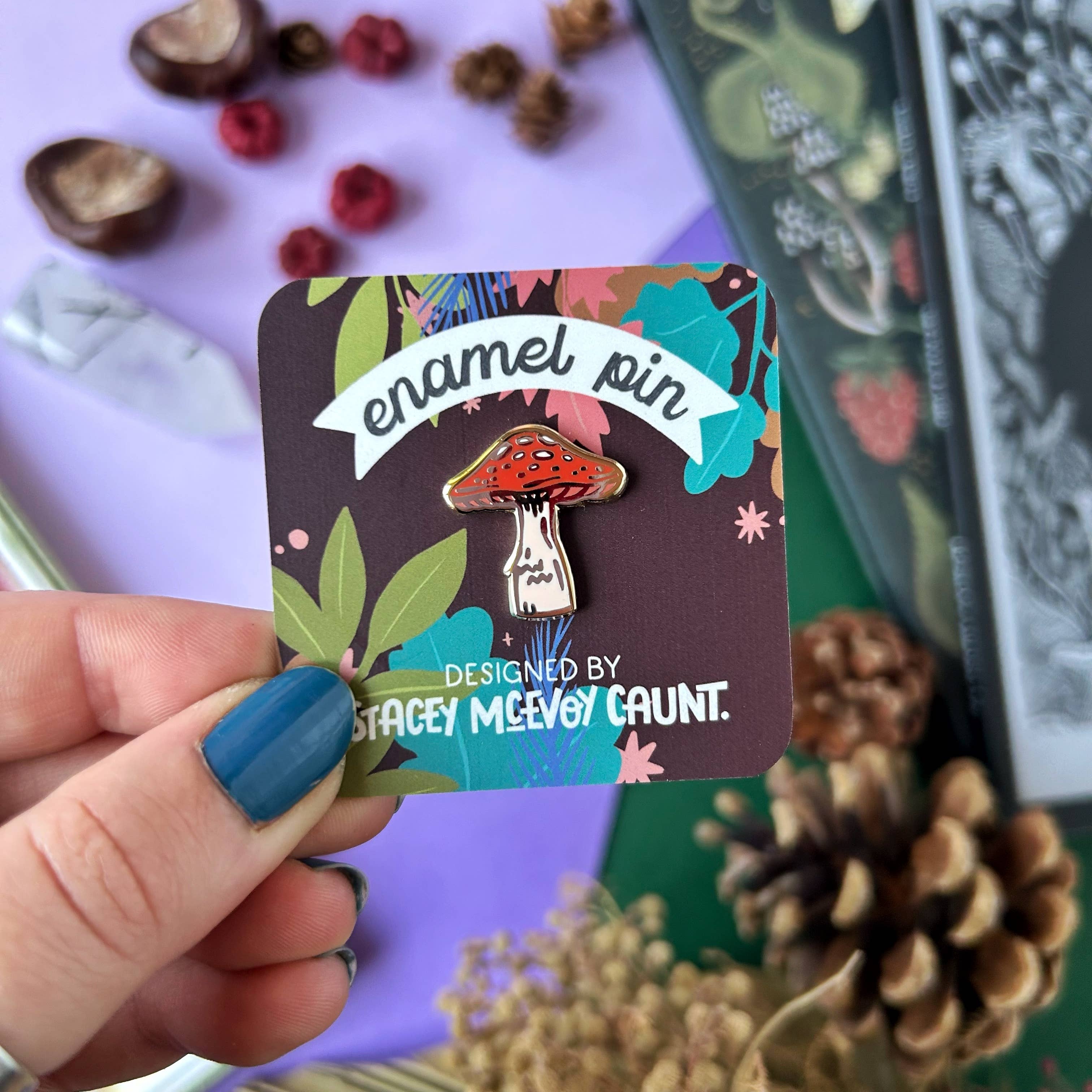 Enamel pin with mushroom design held on a card, surrounded by nature-themed items.