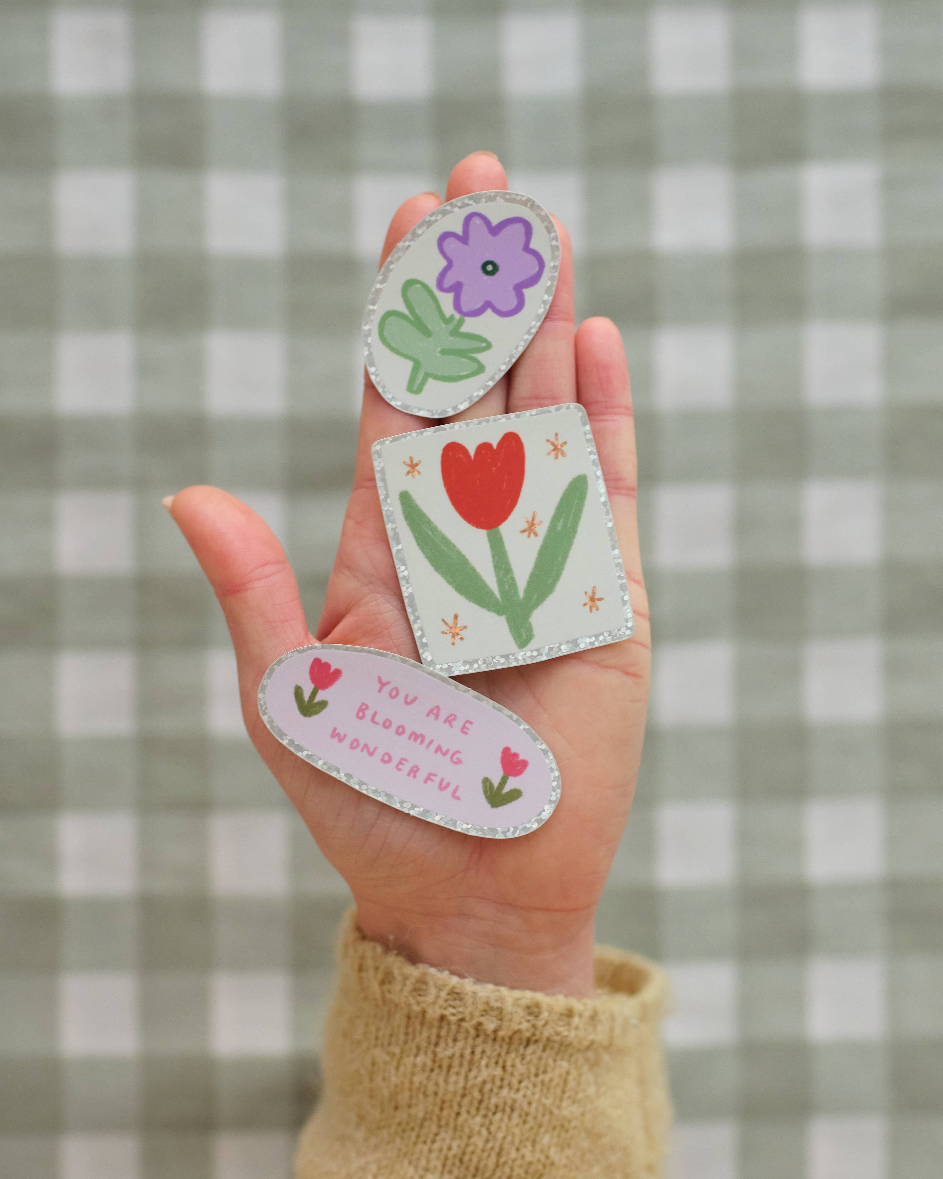 Hand holding three colourful floral stickers with different designs, against a checkered background.