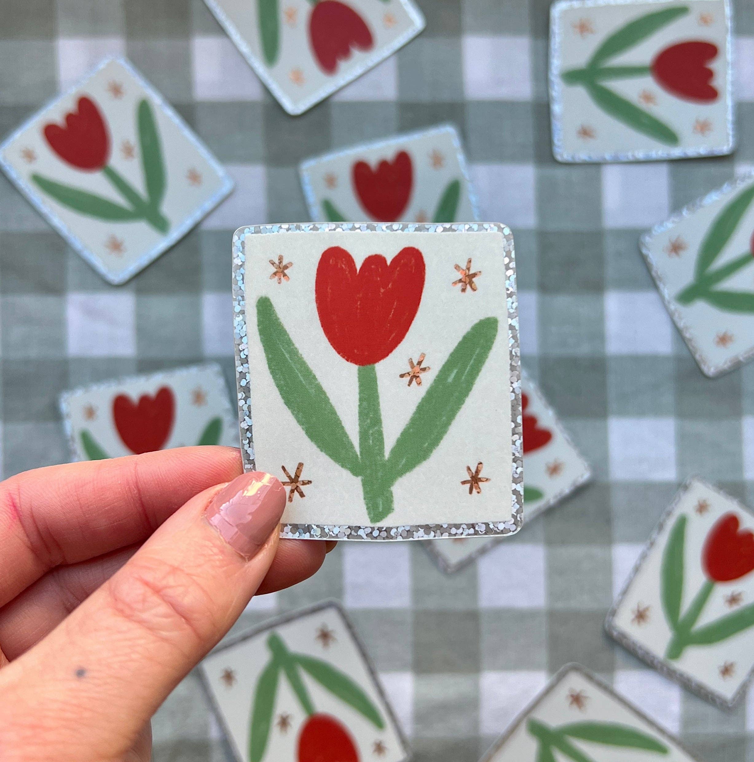 Hand holding a small square sticker with a red tulip design on a checkered background.