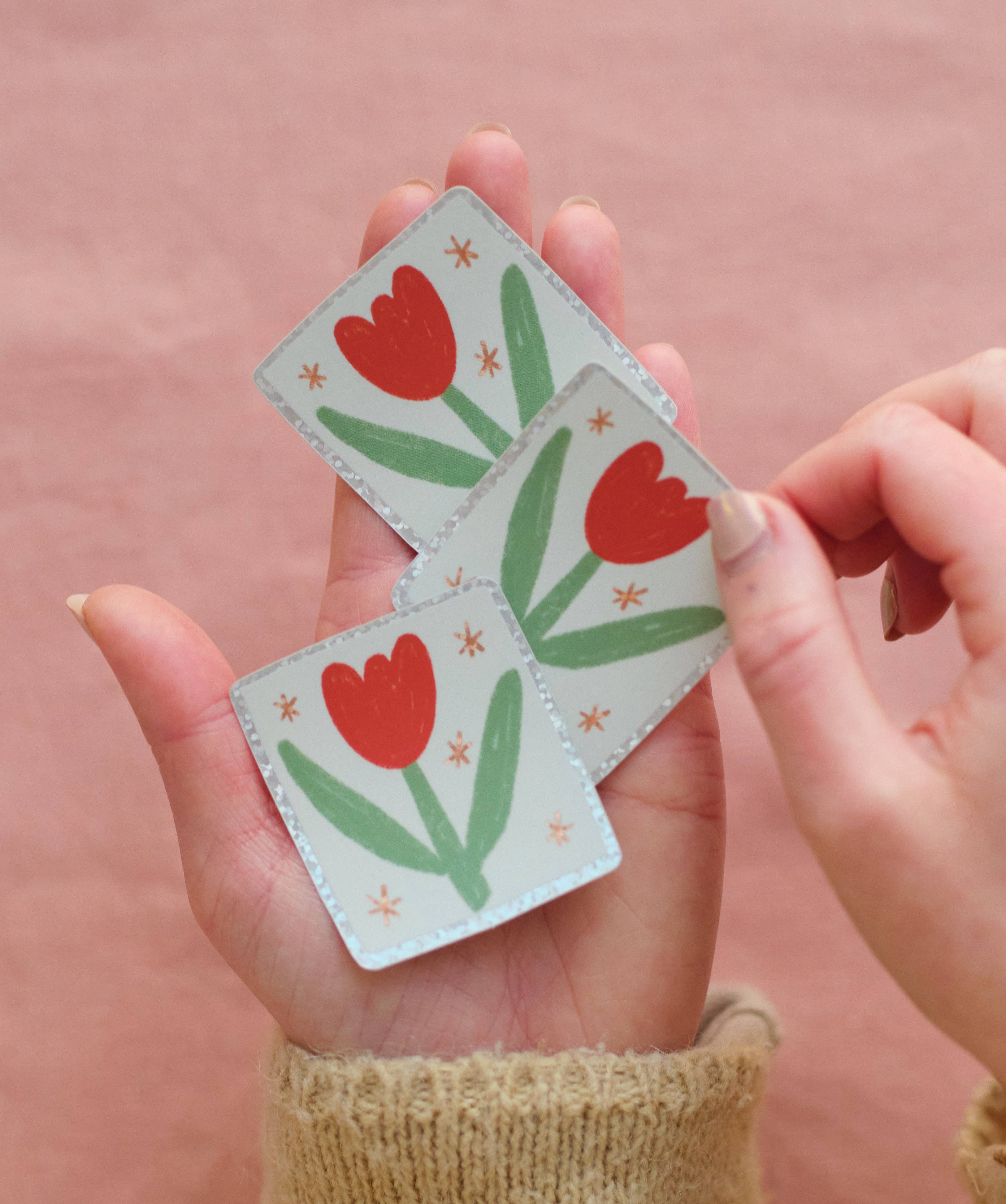 Hand holding three small stickers with red tulip designs on a pink background.