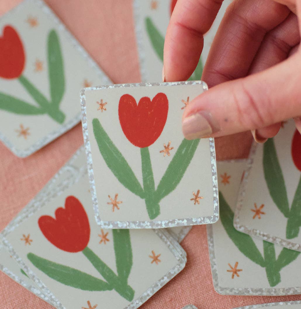 Hand holding a square sticker with red tulip design on a pink fabric background with the same stickers scattered around.