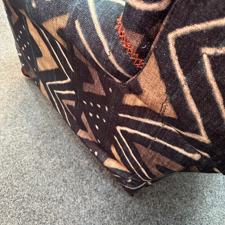 A close-up of a chair featuring a dark and tan geometric pattern with white lines and orange stitching, on a grey carpet.