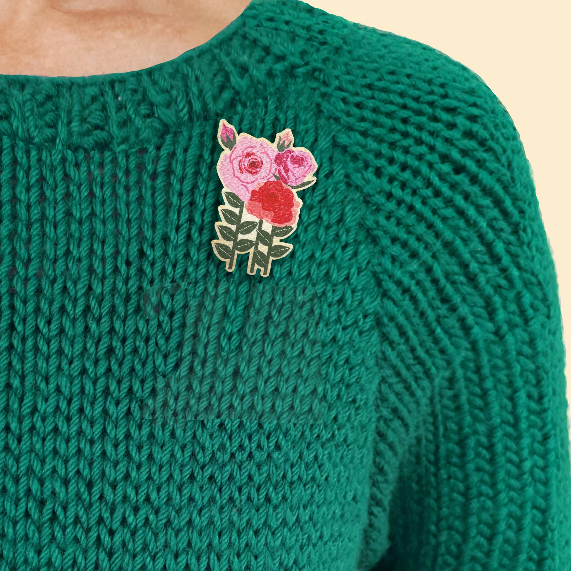 A close-up of a person wearing a green knit sweater and a small floral brooch on the chest. The pin features pink and red roses with green leaves on a cream background.