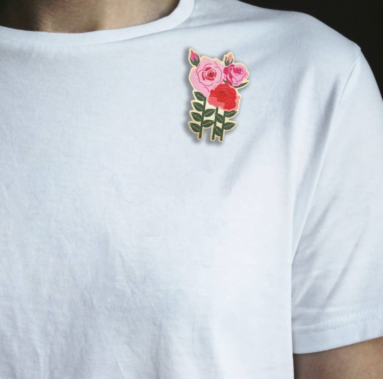 A close-up of a person wearing a white crew-neck t-shirt with a small floral brooch on the left chest. The pin features pink and red roses with green leaves.