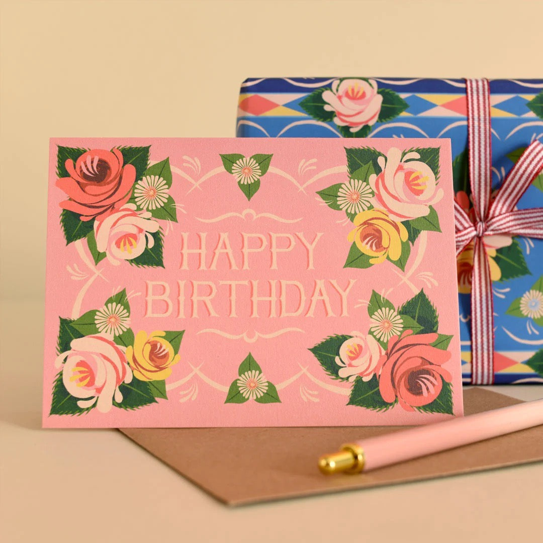 Pink birthday card with floral design and a wrapped gift in the background.