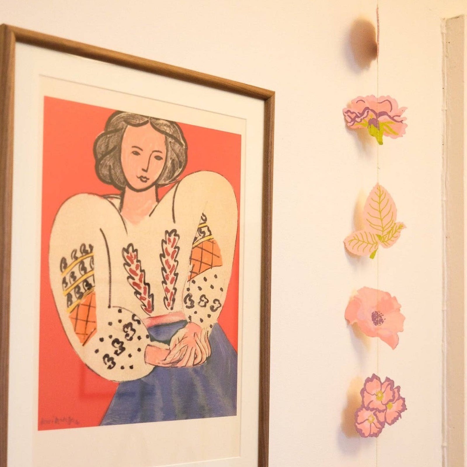 A vertical hanging garland with rosed in shades of pink and purple and green leaves hanging next to a print of a lady in a white shirt.