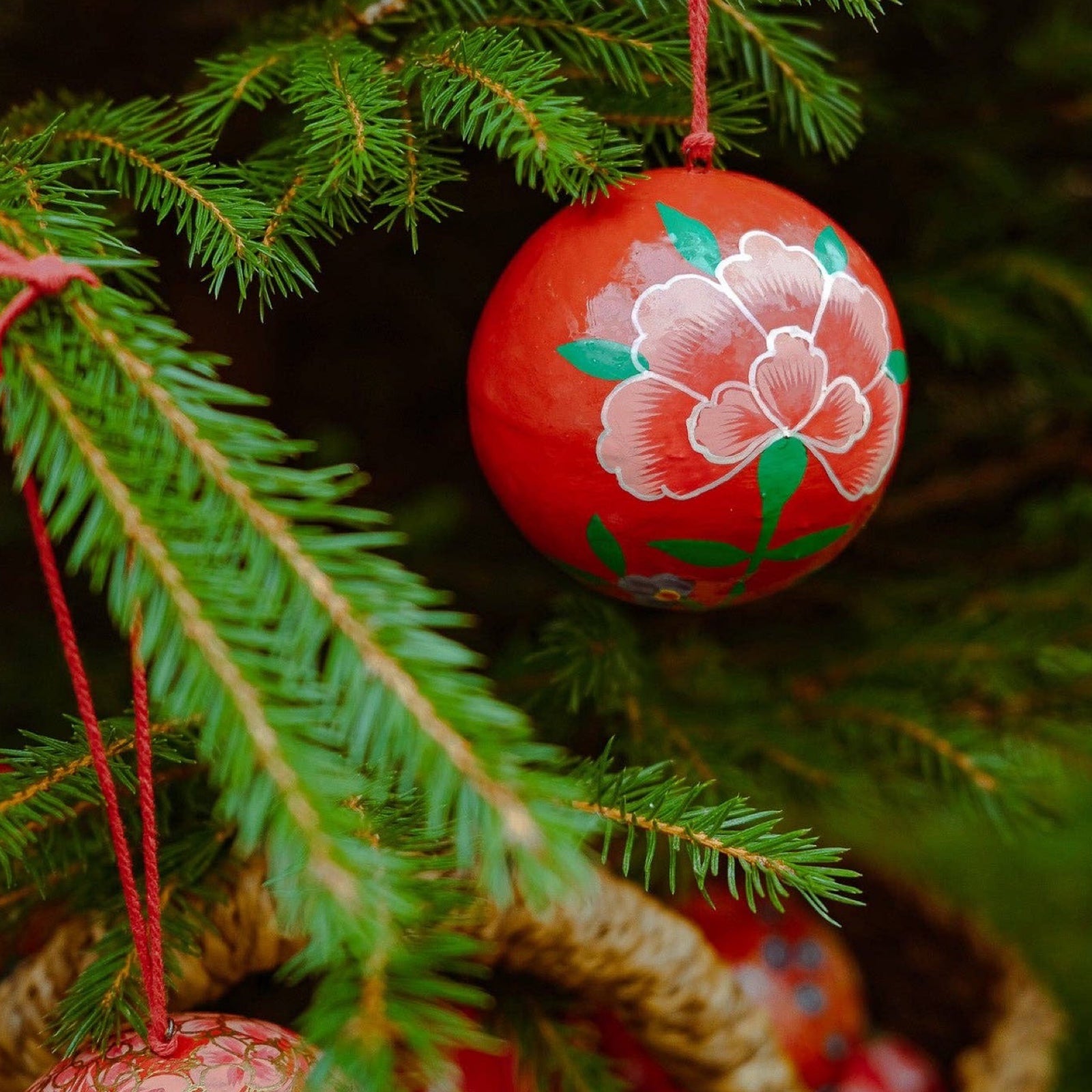 A round papier mache bauble in red with a red, green and white floral design hanging on a Christmas tree.