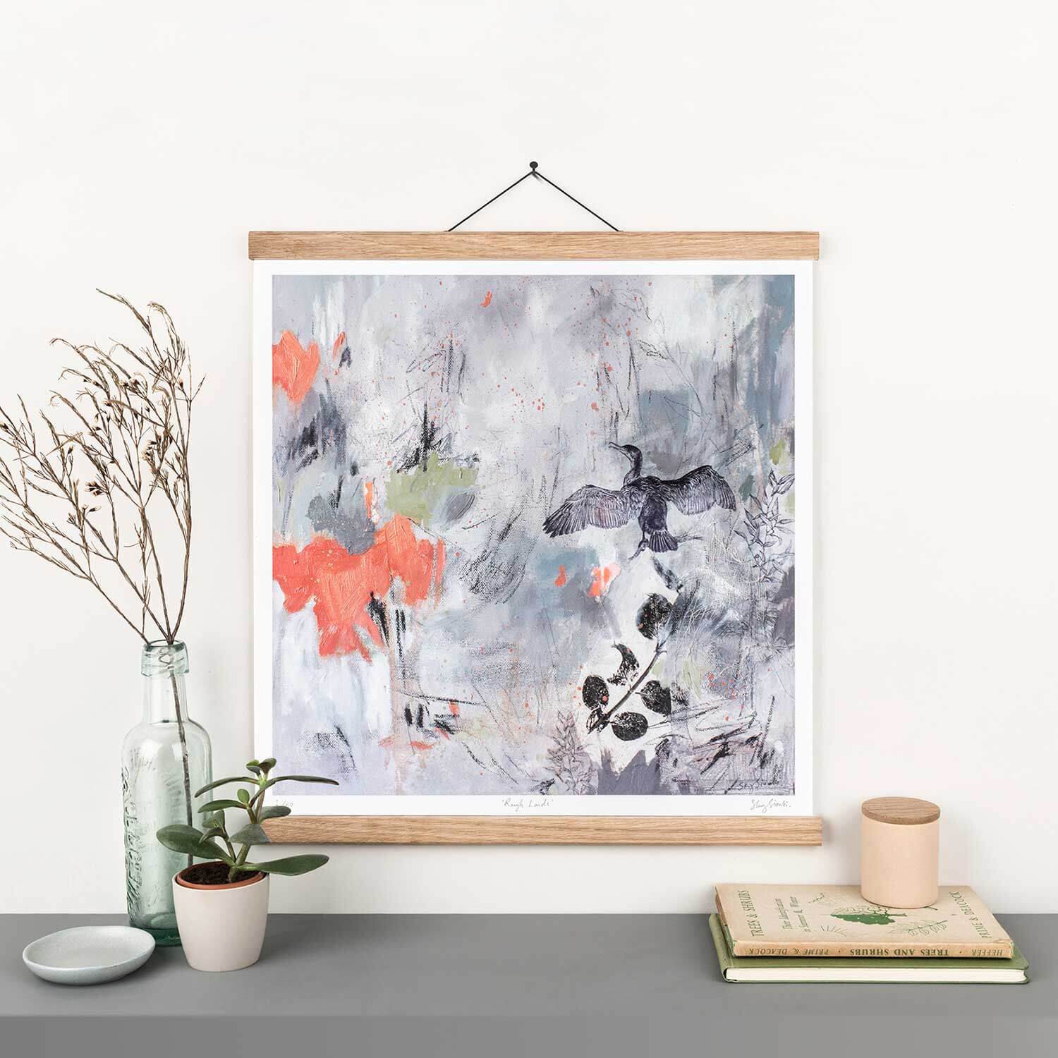 A framed bird print featuring hand-drawn illustration of a bird in flight with botanical leaves, in shades of grey, green and orange, displayed on a white wall.