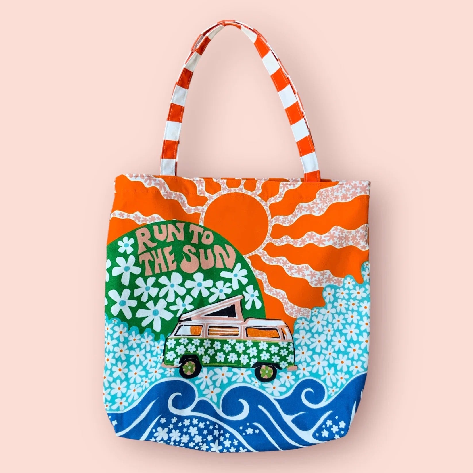 A fabulous, bright and colourful tote bag featuring a pink campervan covered in green daisies on a meadow of blue daisies, a large orange sun and blue waves.