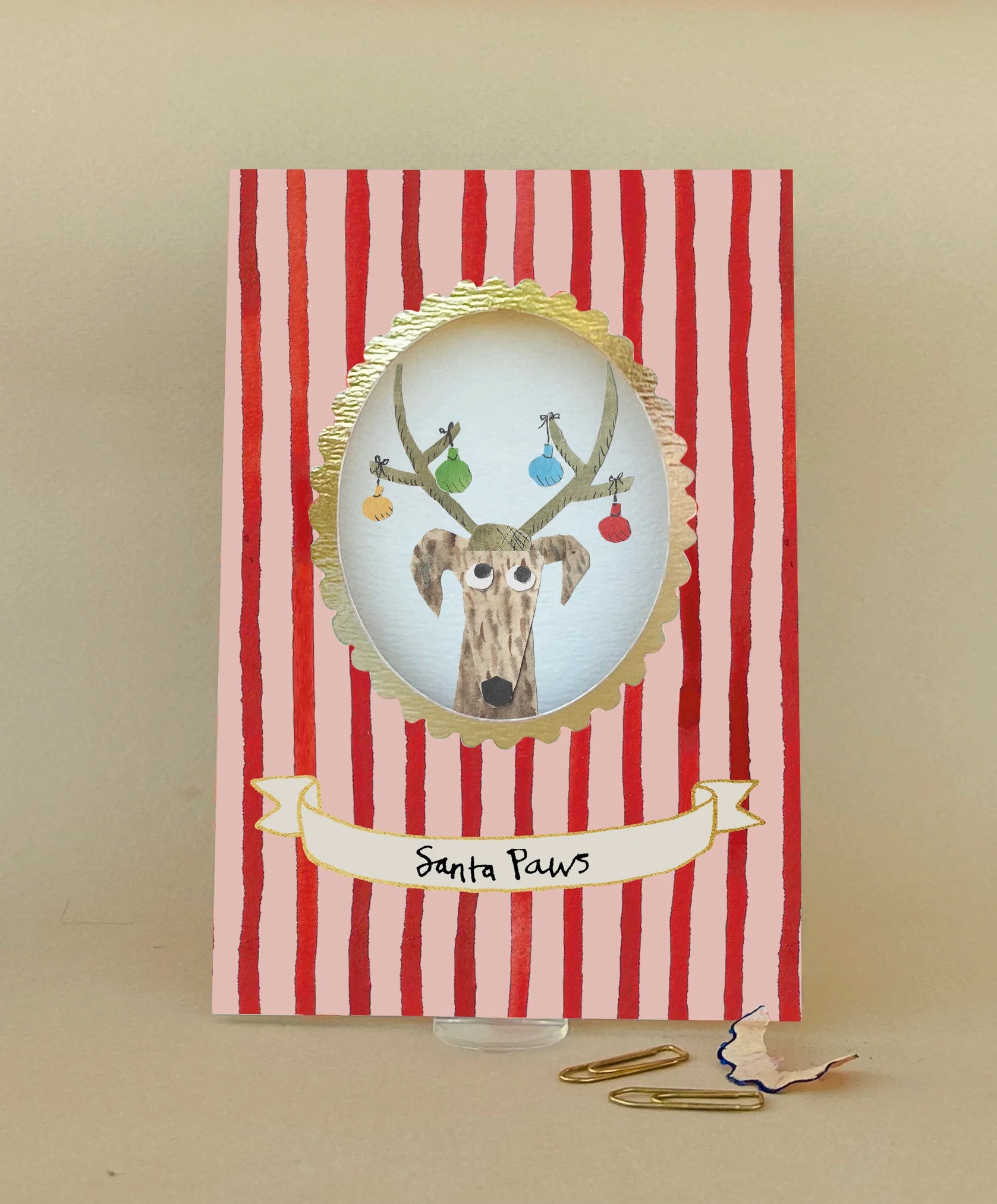 A Christmas greeting card with red and white vertical stripes, featuring an oval with an illustration of a dog with antlers wearing ornaments, and a banner reading 'Santa Paws'.