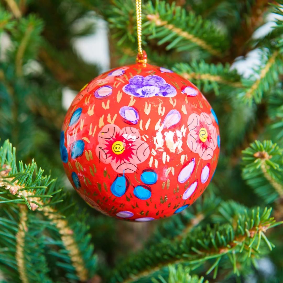 A round papier mache bauble in red with a lilac, blue and red floral design hanging on a Christmas tree.