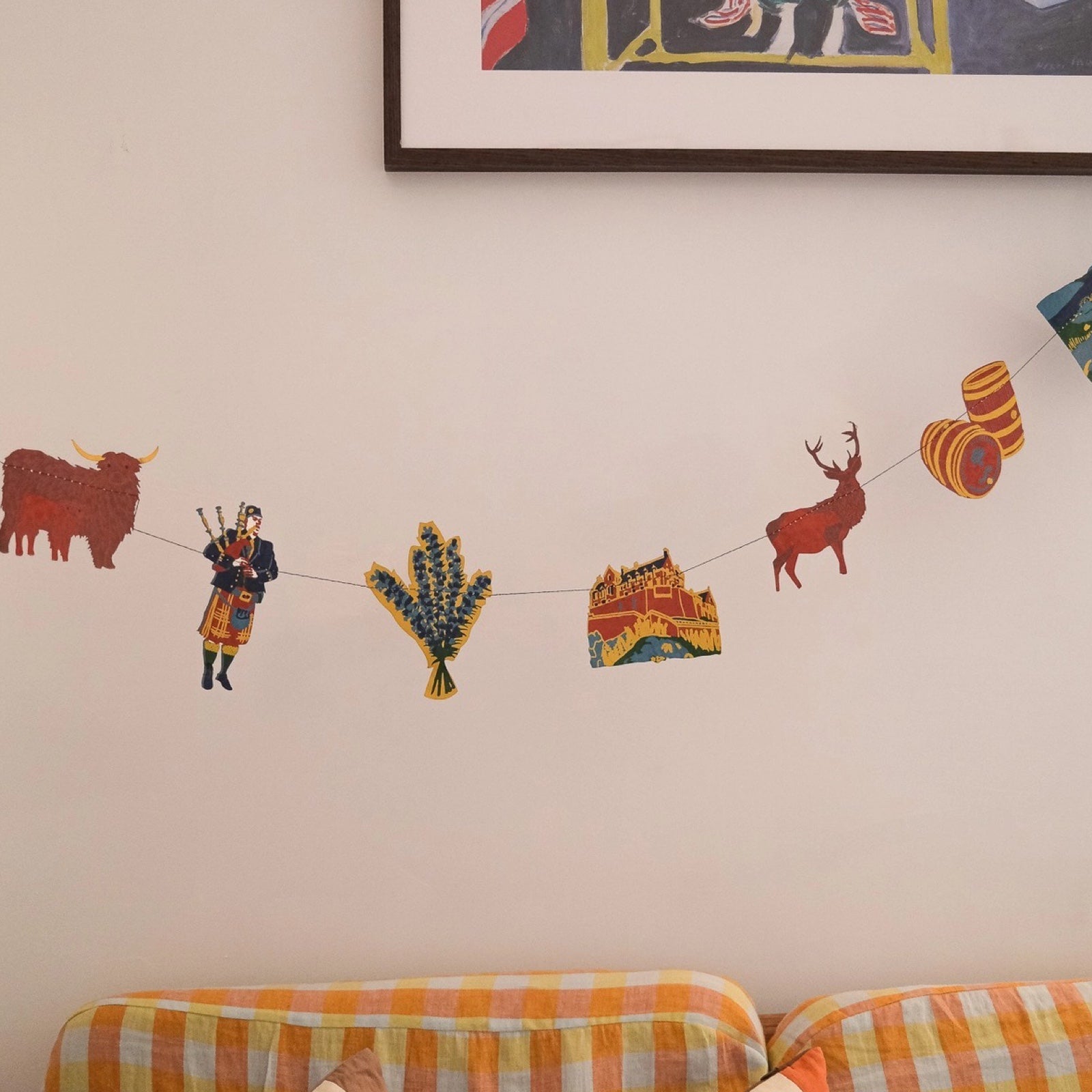 A Scotland paper garland featuring fourteen screen-printed paper Scottish-themed designs (pipers, scottie dogs, Edinburgh Castle, stags and thistles hanging on a cream wall above an orange checked sofa.