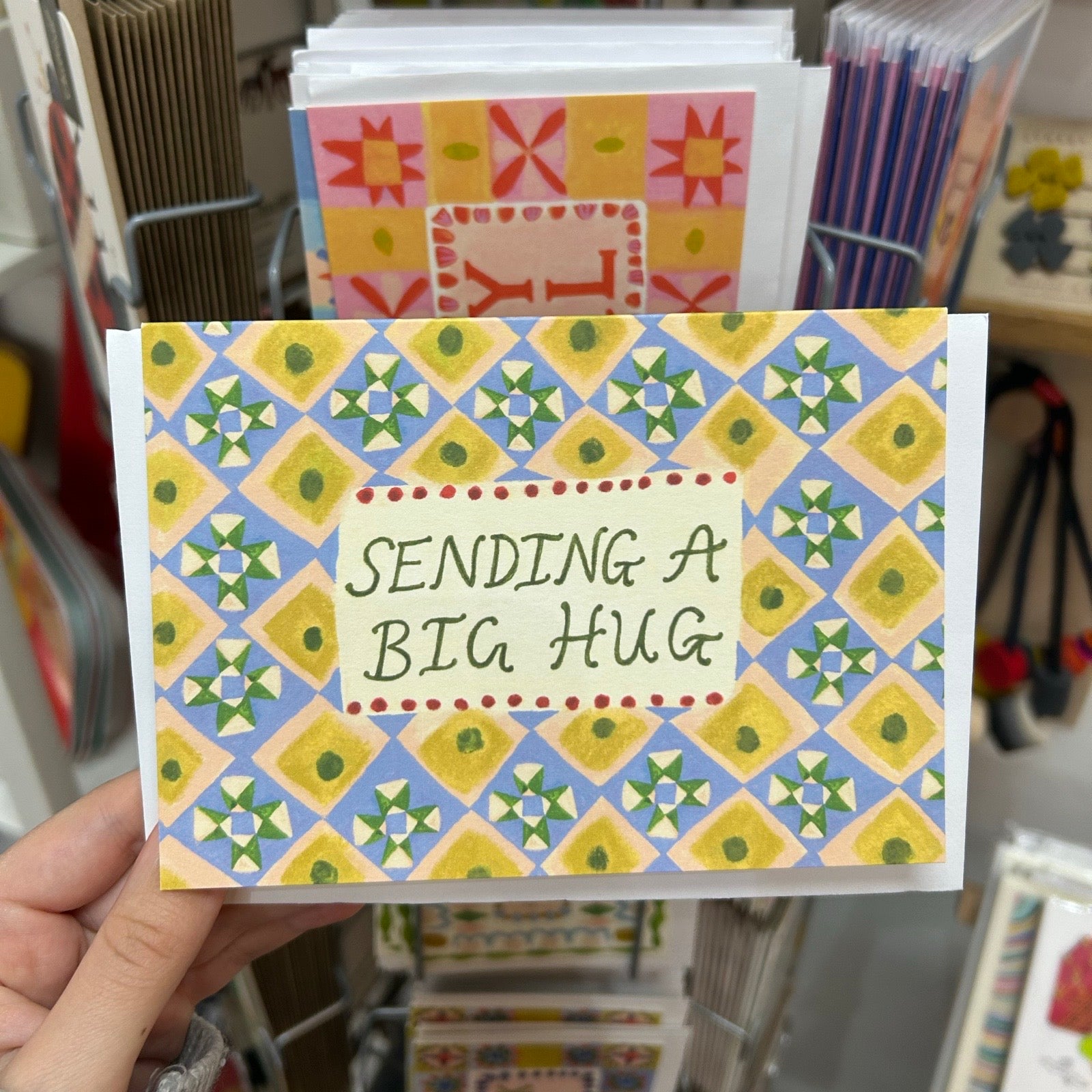 Card with geometric pattern and 'Sending a Big Hug' text held in front of a display of greeting cards.