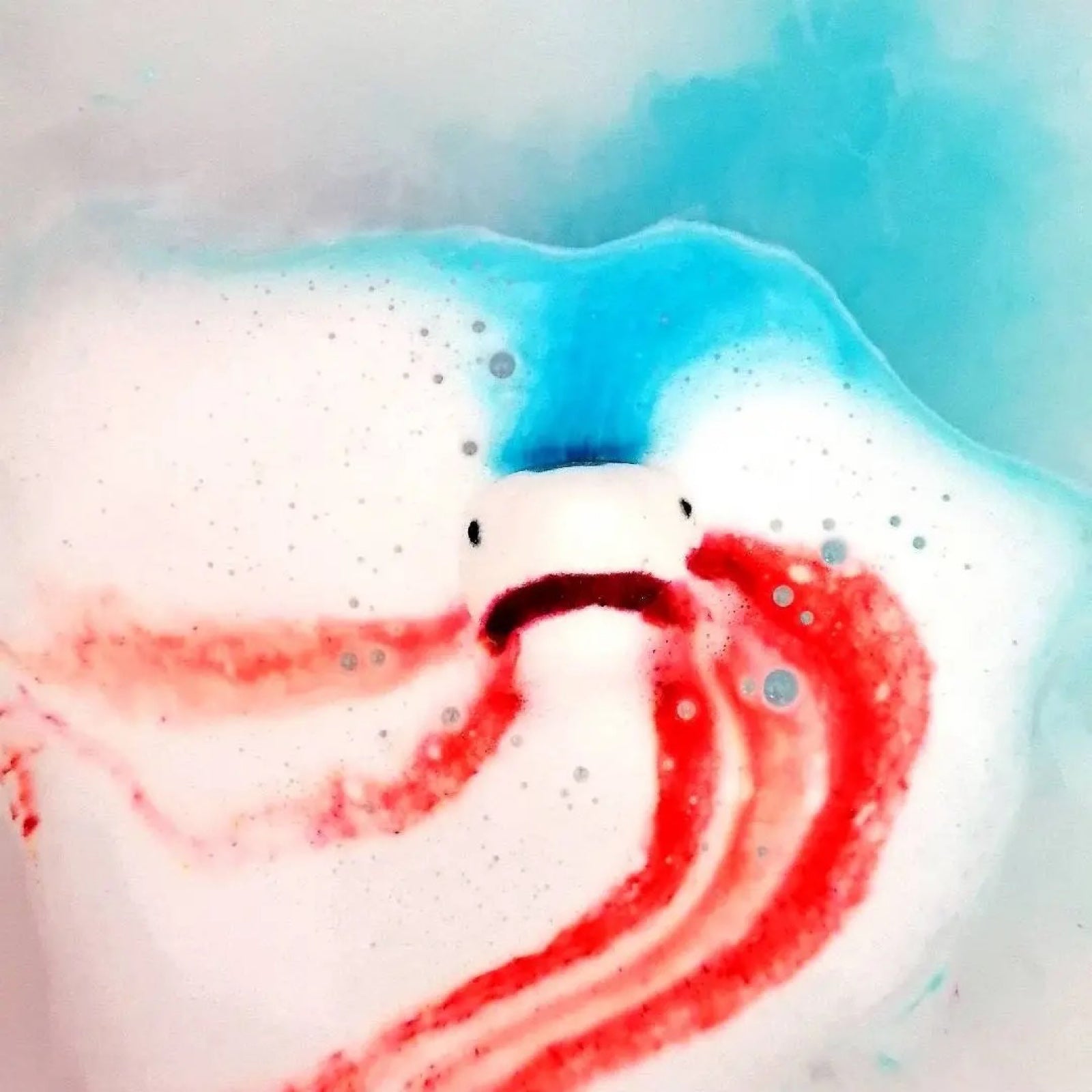 A shark head shaped bath bomb dissolving in the bath and turning the water blue, white and red.