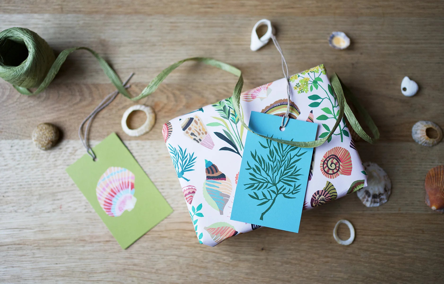 Colourful gift tags with nature-themed designs on a present in shell wrapping on a wooden surface with twine and shells.