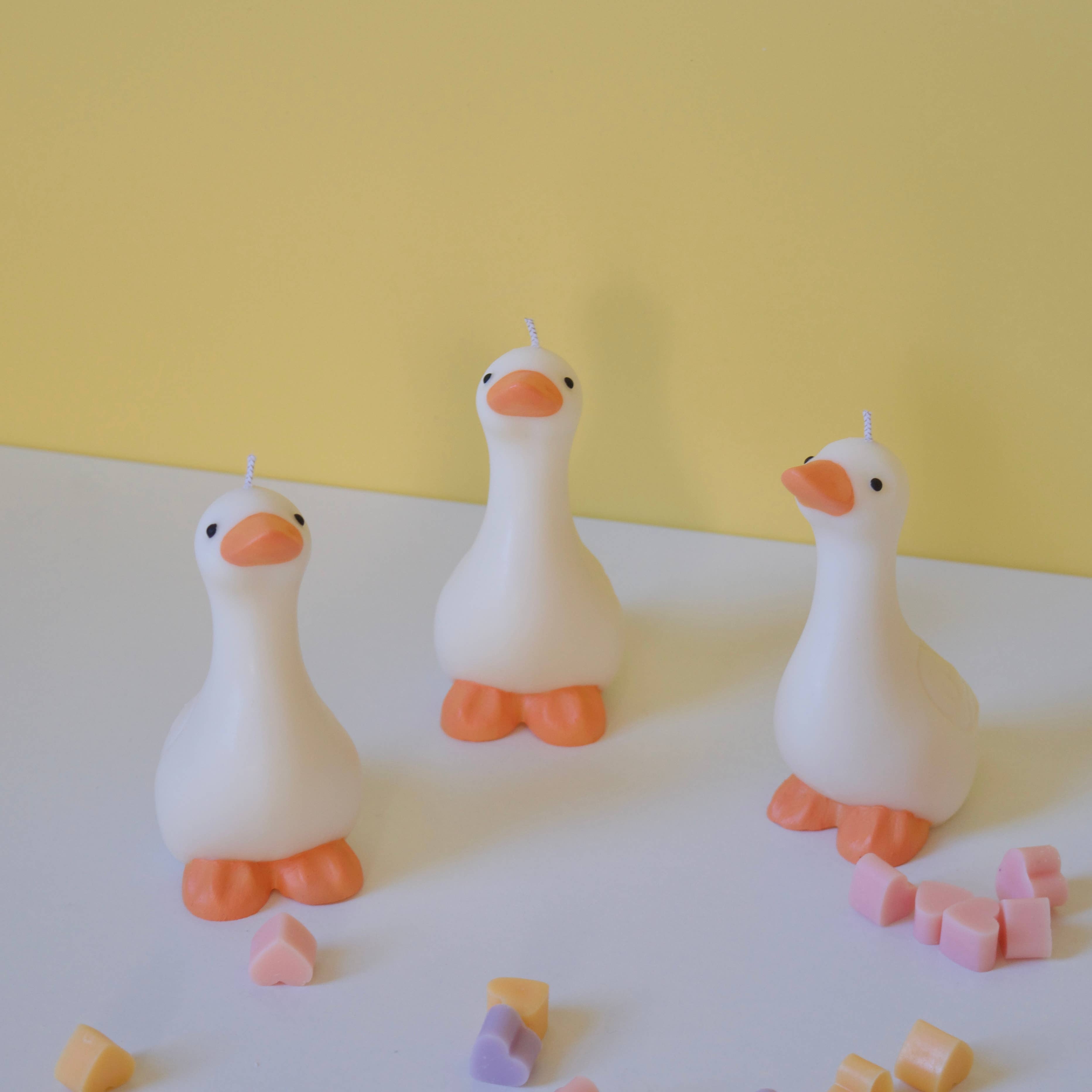 Three white goose-shaped candles with orange beaks and feet on a white surface with a yellow background.