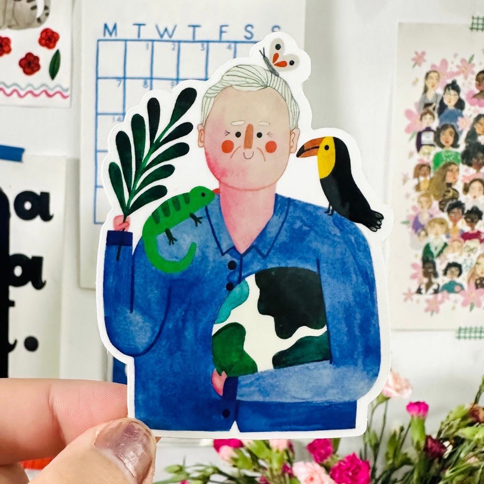 Illustrated sticker of David Attenborough with plants and animals, held by a hand, with a decorative background.
