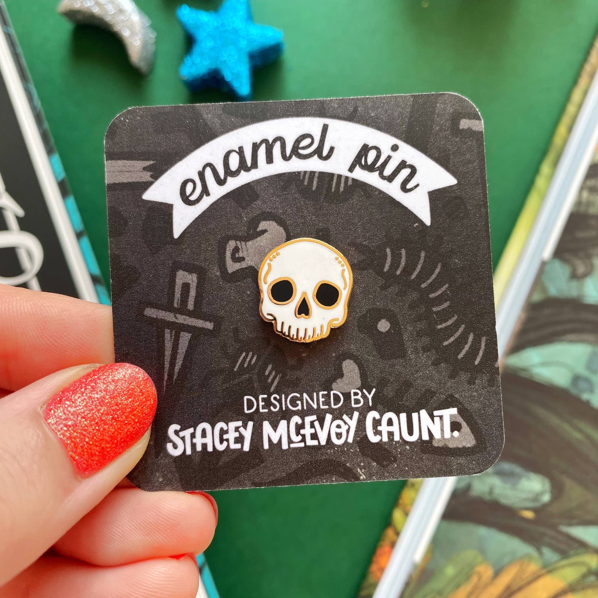 Enamel pin with skull design held on a card labeled 'Designed by Stacey McEvoy Caunt'.