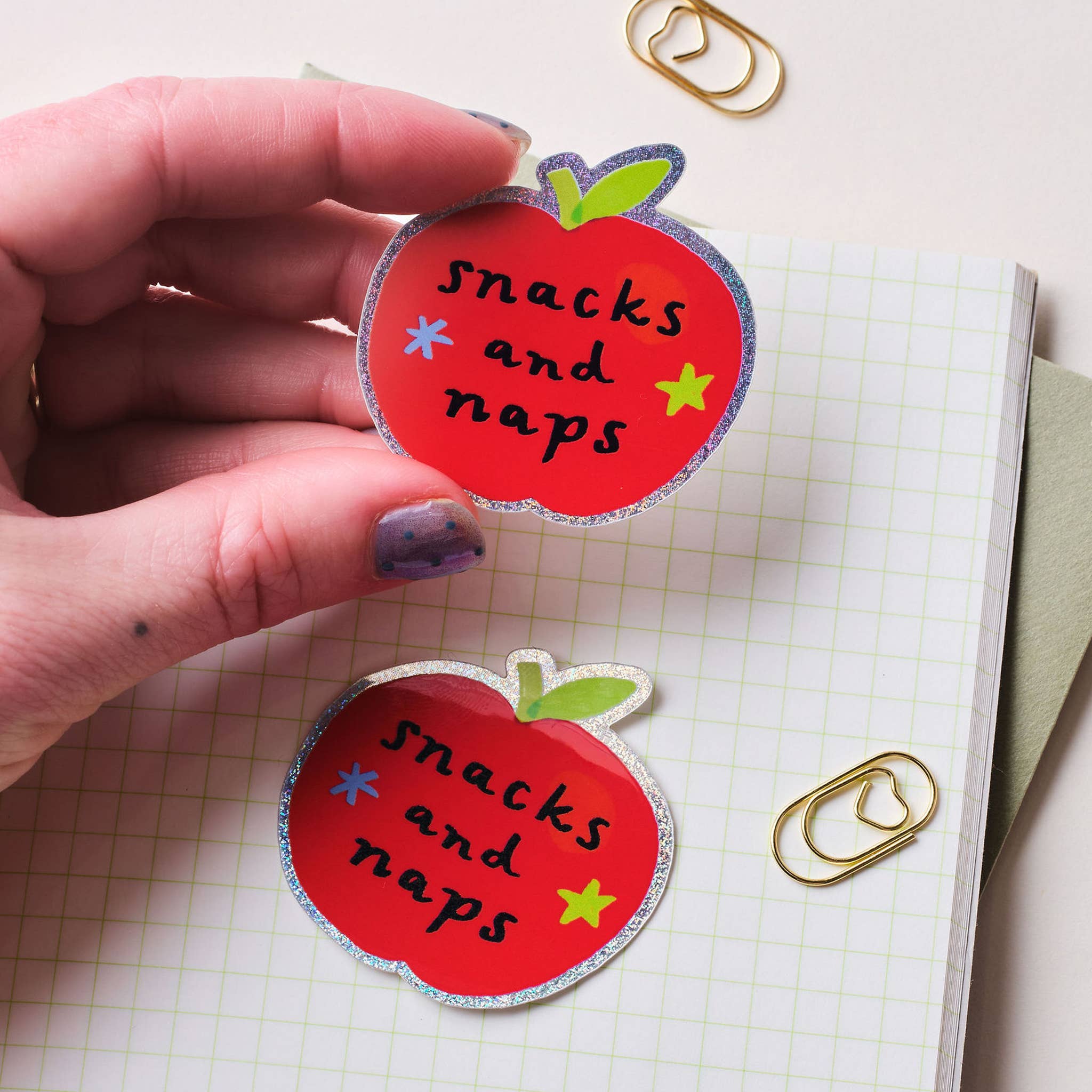 Hand holding a red apple-shaped sticker with 'snacks and naps' text on a checkered background.