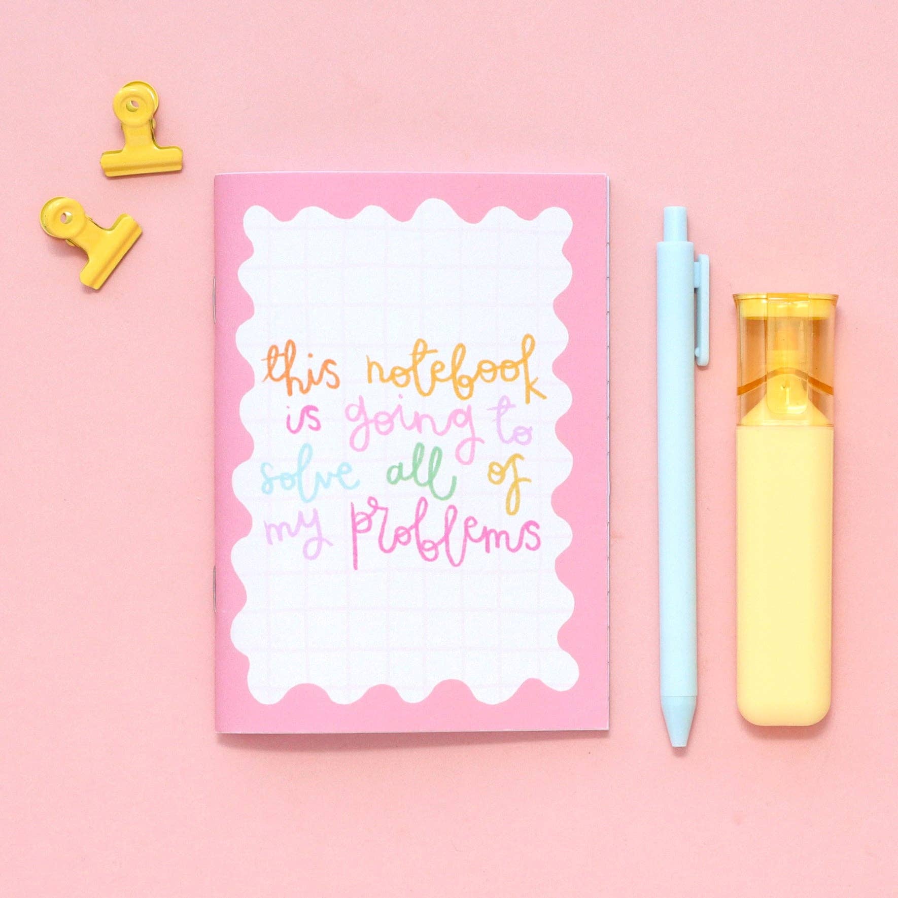Pink notebook with colourful text, light blue and yellow pens, yellow clips on a pink background.
