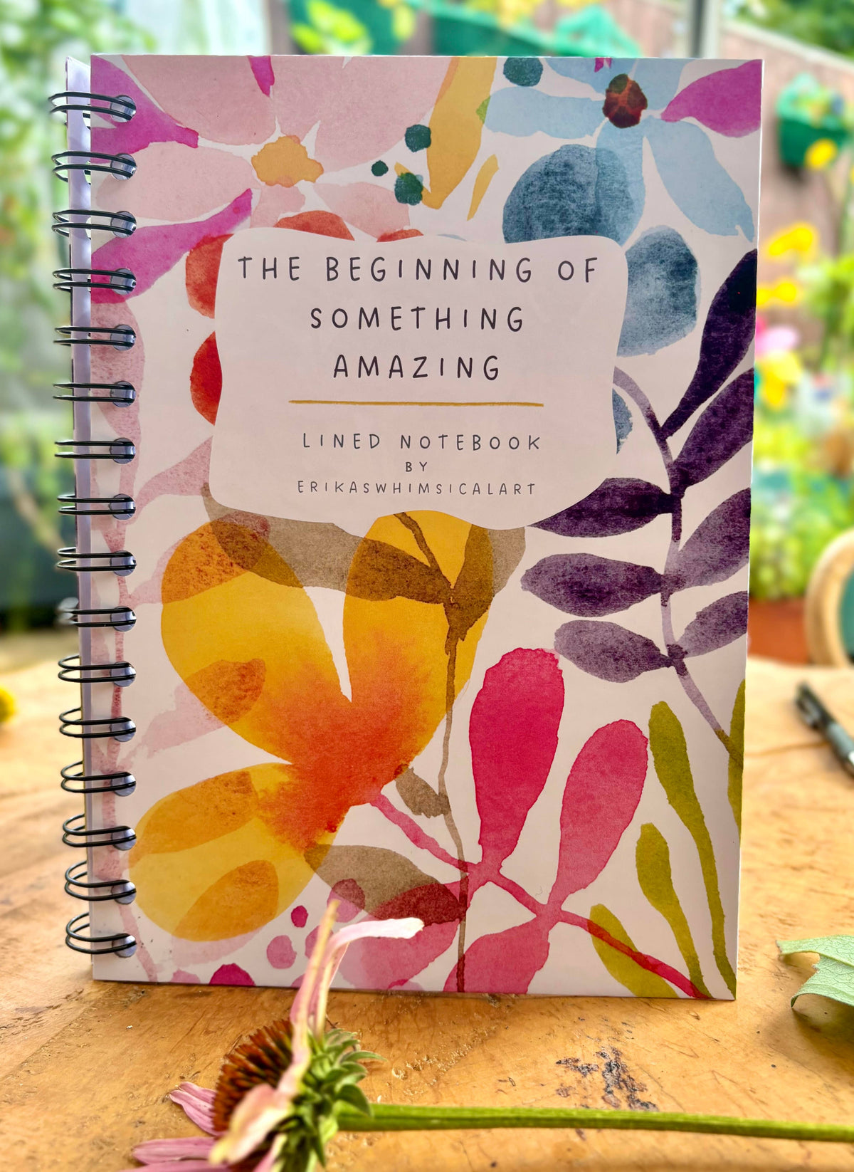 A spiral-bound lined notebook titled 'The Beginning of Something Amazing' by Erika Whimsical Art, featuring a colorful watercolor floral cover, displayed on a wooden surface with a blurred garden background.