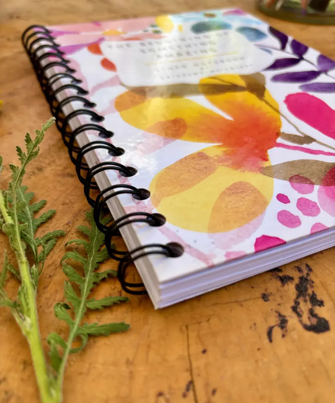 A spiral-bound notebook with a colourful watercolor floral cover lies on a wooden tabletop next to a green herb stem. The cover has a white base with pink, yellow, orange, purple, and blue abstract floral patterns, and a small white label near the top.