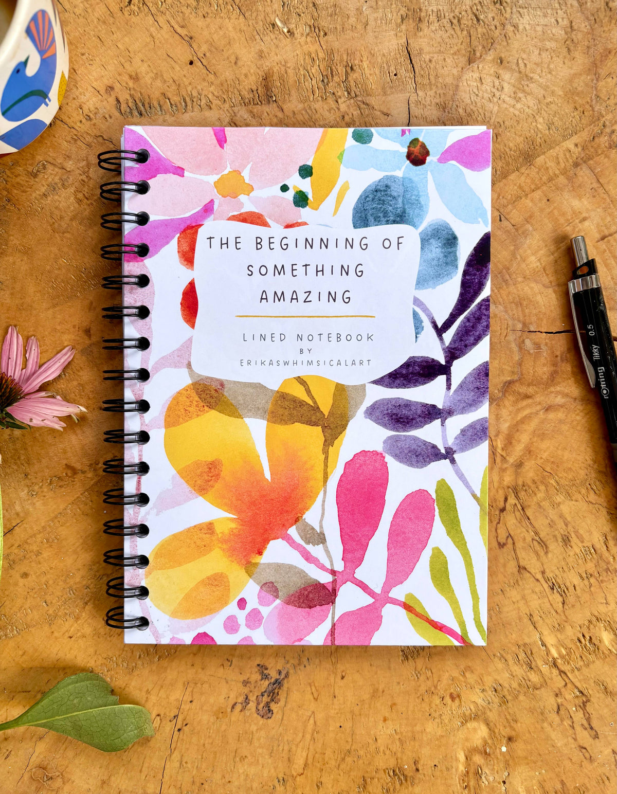 A spiral-bound lined notebook with a colorful watercolor floral cover sits on a wooden tabletop. The cover reads 'THE BEGINNING OF SOMETHING AMAZING' and 'LINED NOTEBOOK'.