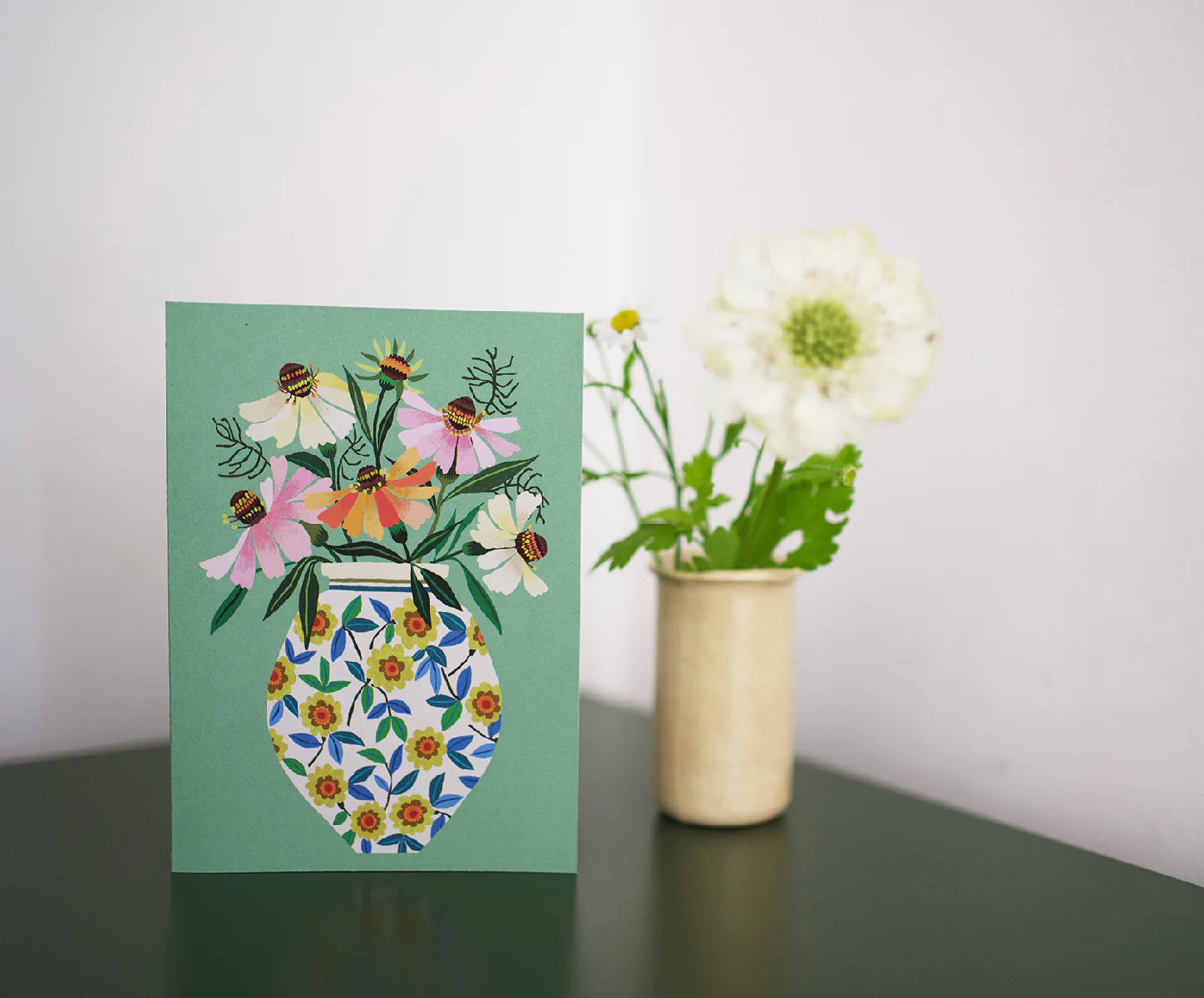 Greeting card with floral design on a green background, placed next to a vase with flowers.