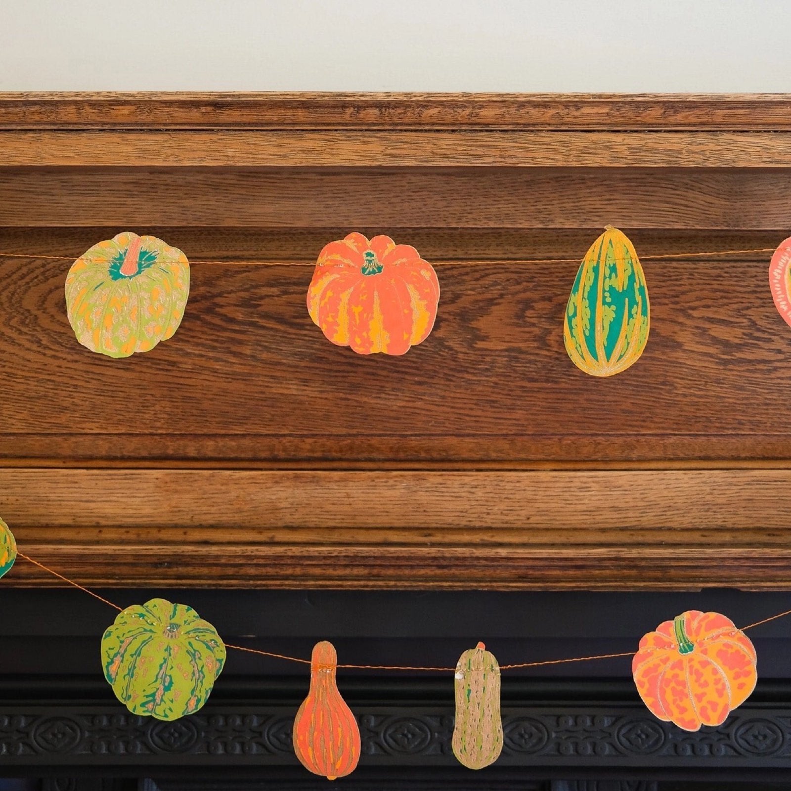 Squashes paper garland, featuring twenty screen-printed pumpkins, squashes and gourds in shades of orange and green hanging on a wooden fireplace.