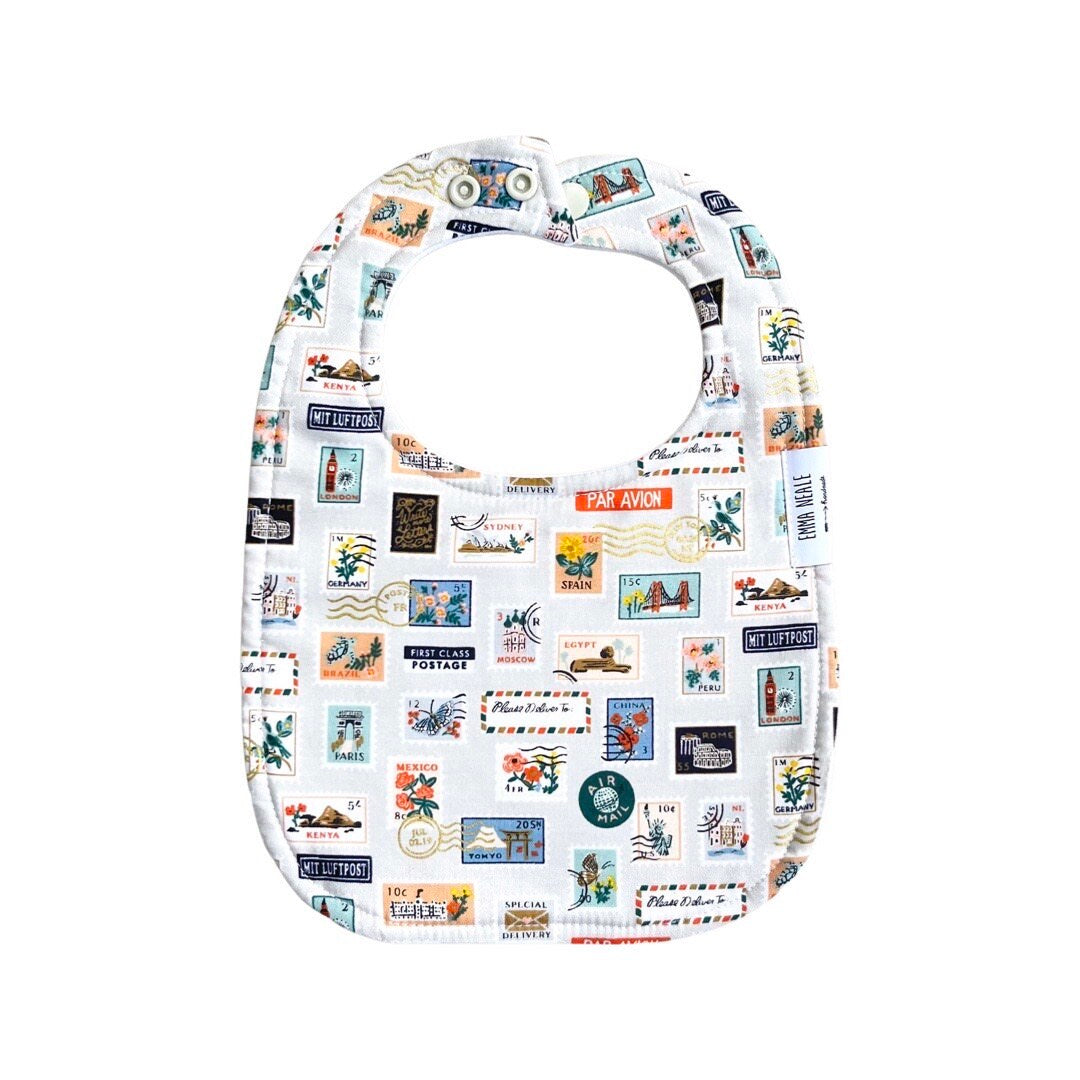 A white baby bib with a colorful pattern of stamps and related imagery, featuring adjustable neck fasteners.