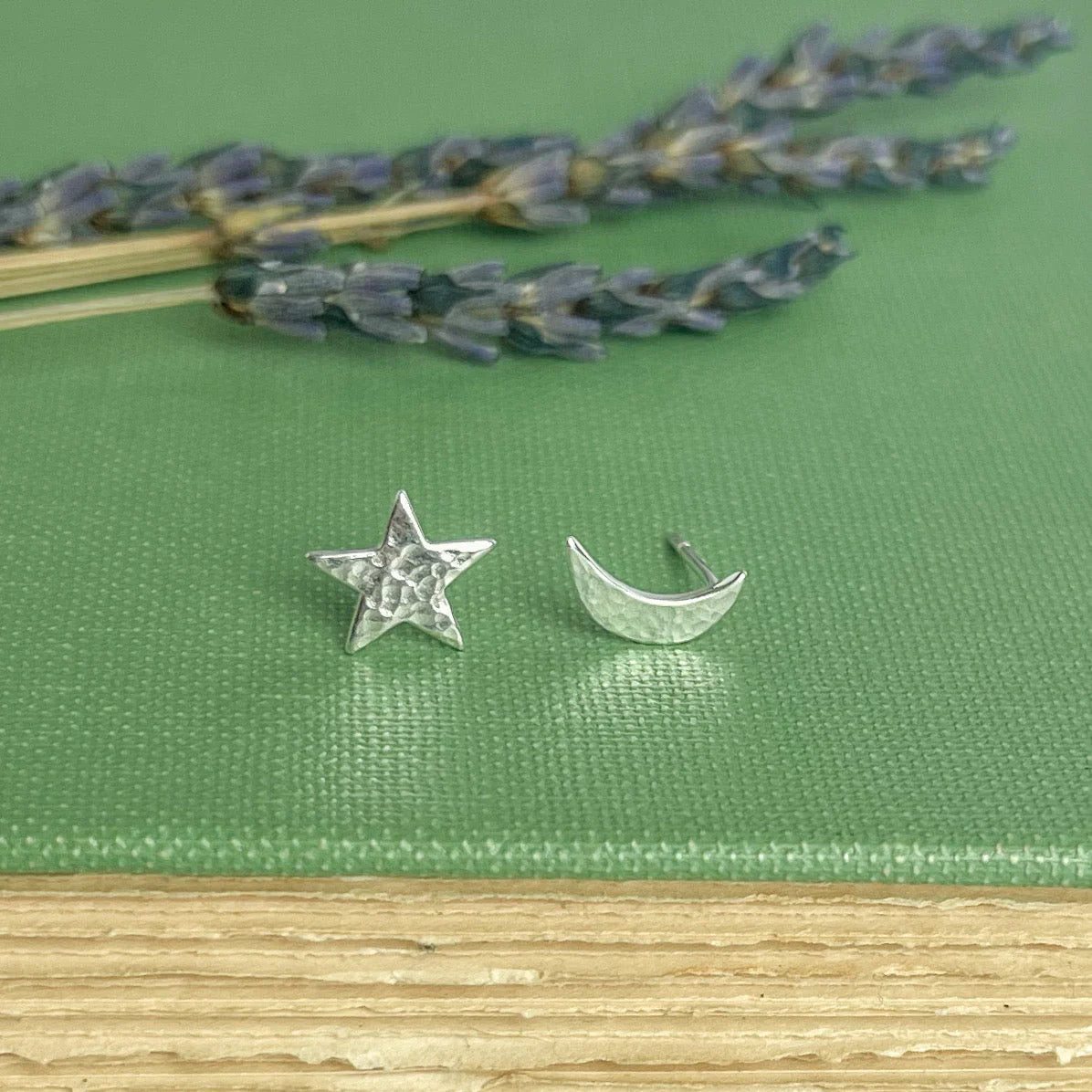 Silver star and moon earrings on a green fabric background with lavender.