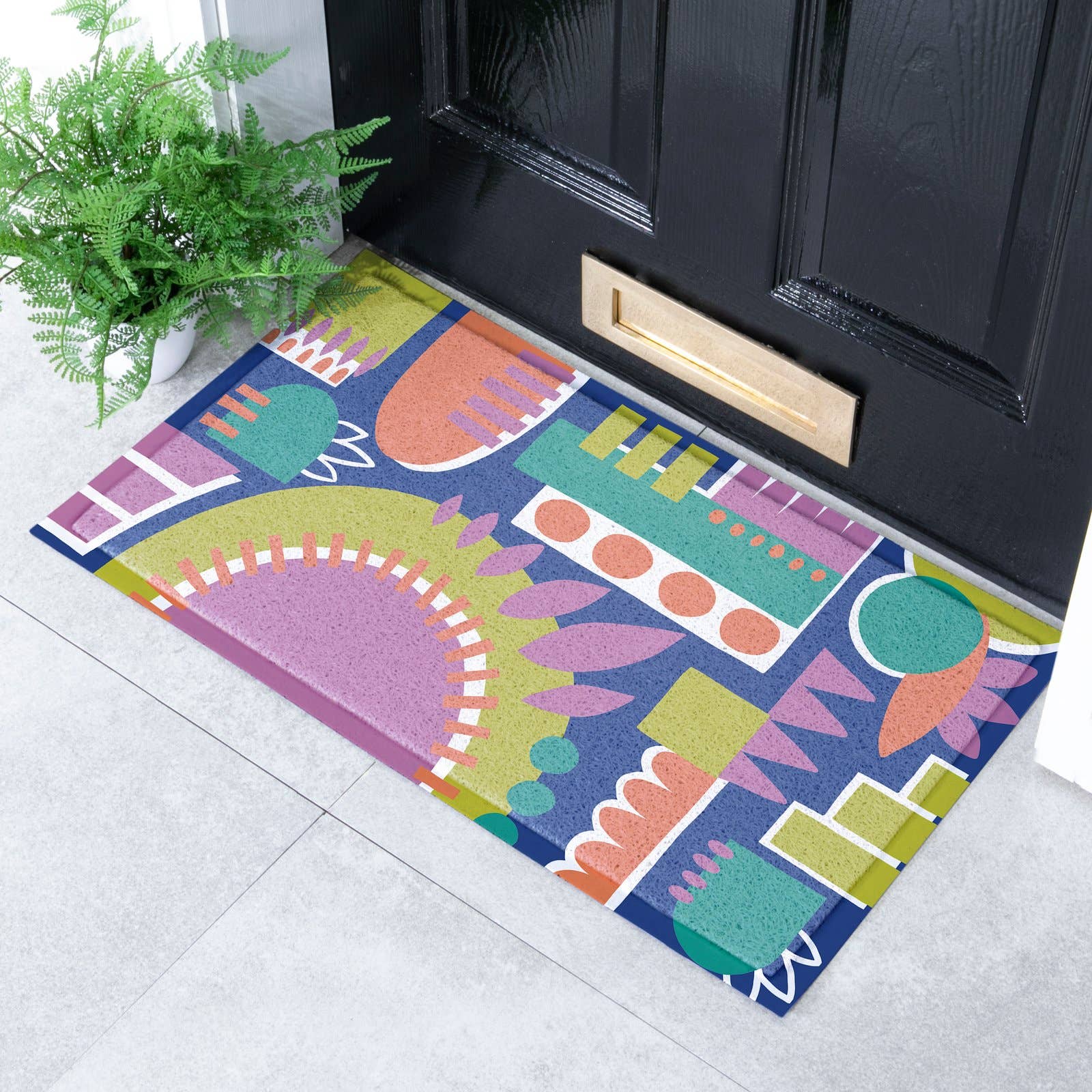A colourful geometric doormat with a modern abstract pattern in shades of purple, green, orange, yellow, and blue, placed in front of a black front door on a light grey tiled floor next to a potted fern.