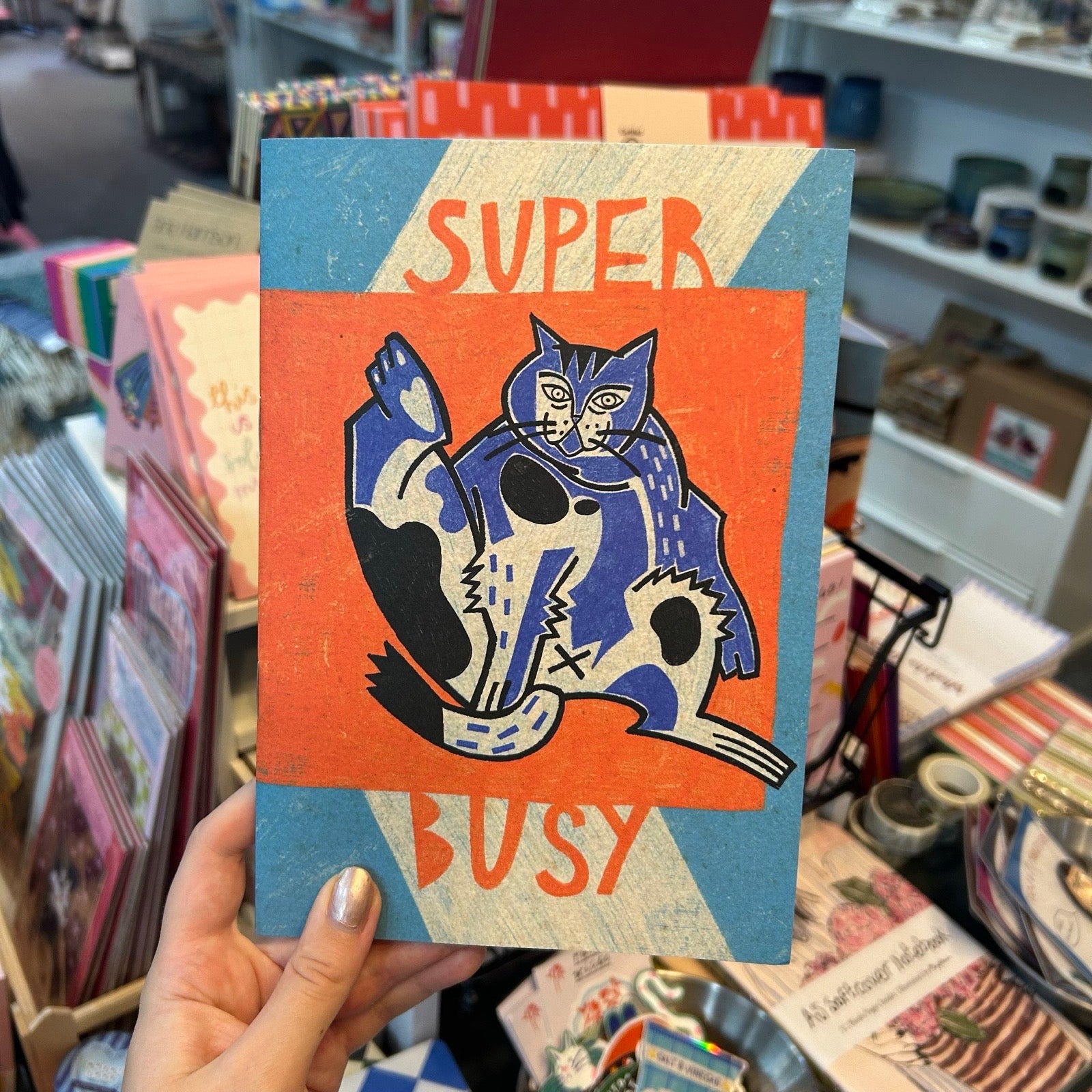 A hand holds a notebook featuring a stylised blue cat on an orange background with the words 'SUPER BUSY' in orange, set against a blurred store background.