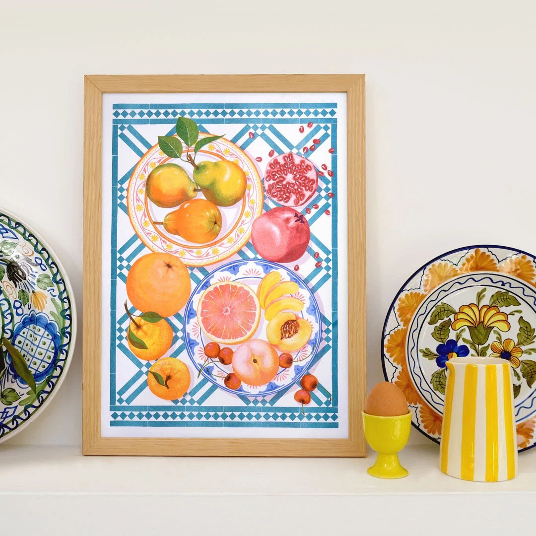 Framed artwork of fruits on a decorative plate with other plates and a vase in the background.