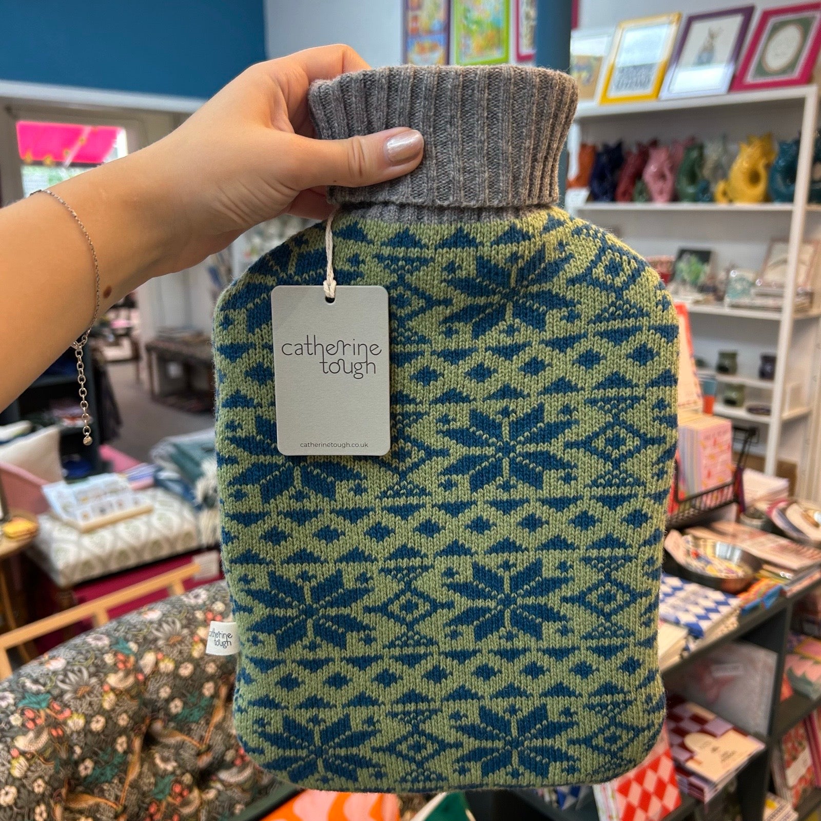 A hand holds a hot water bottle covered in a knitted cover by Catherine Tough. The cover features a blue and sage green geometric snowflake pattern, with a ribbed gray turtleneck.