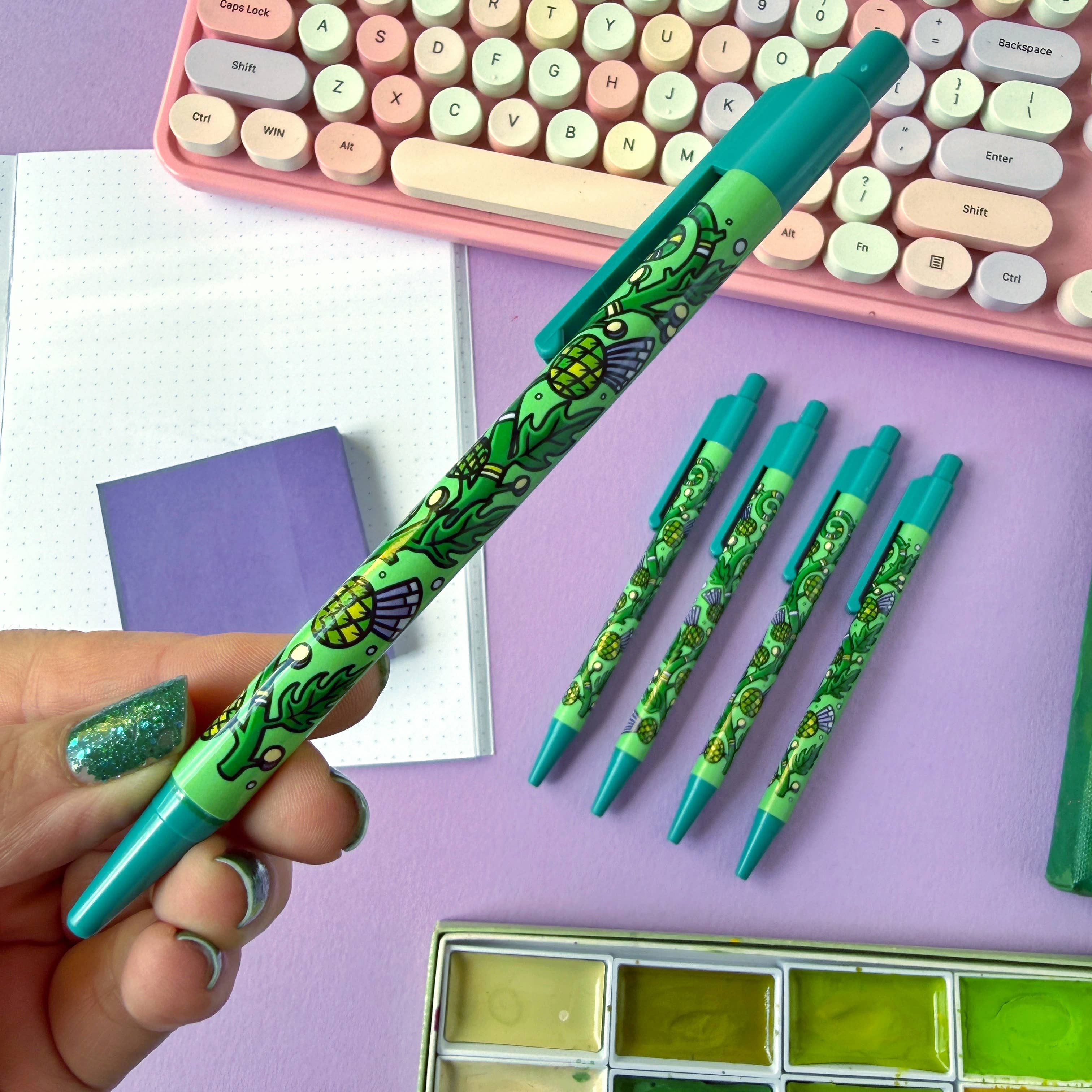 Set of green pens with thistles on a purple surface, with a hand holding one pen.