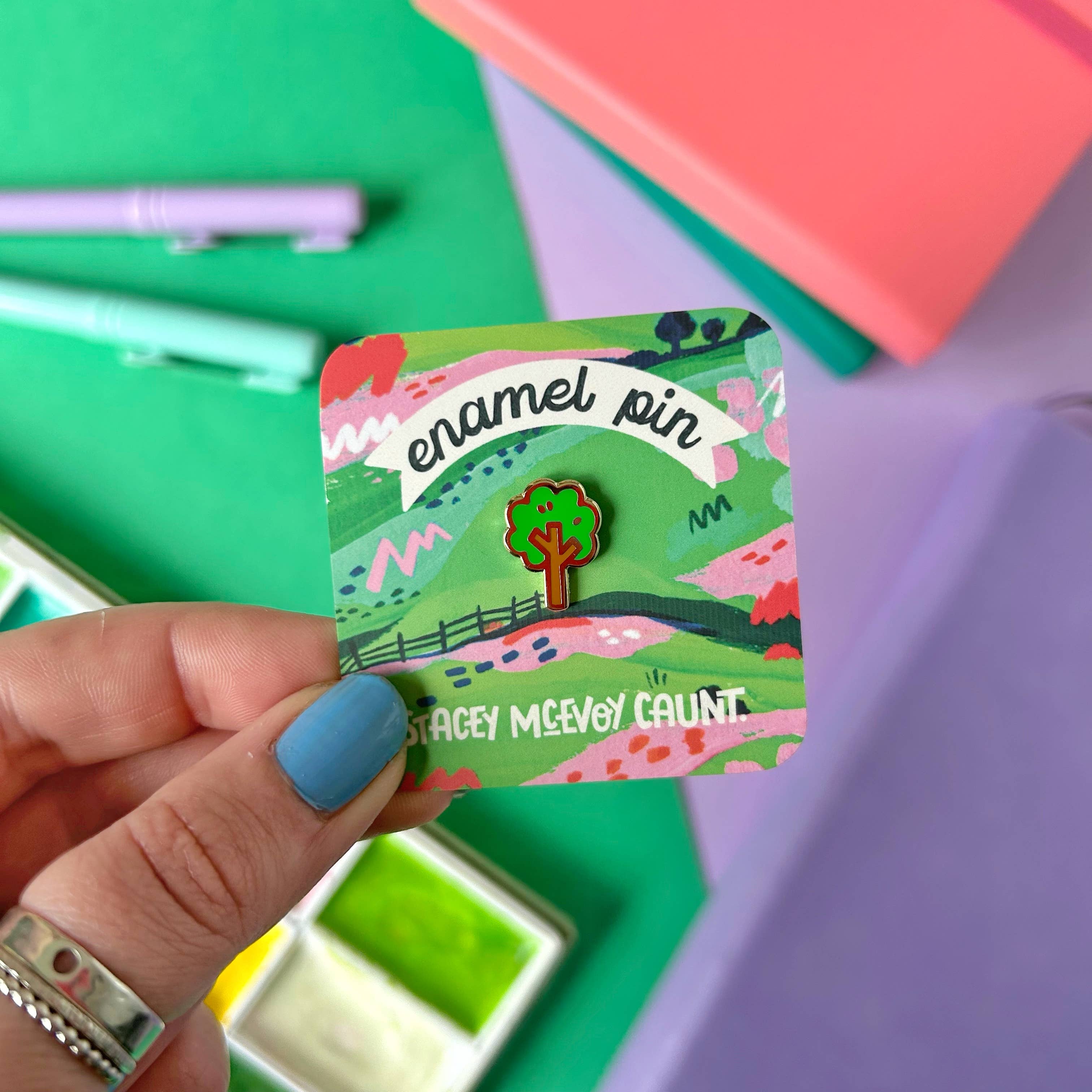 Hand holding a scenic enamel pin with a colourful card against a multicoloured background.