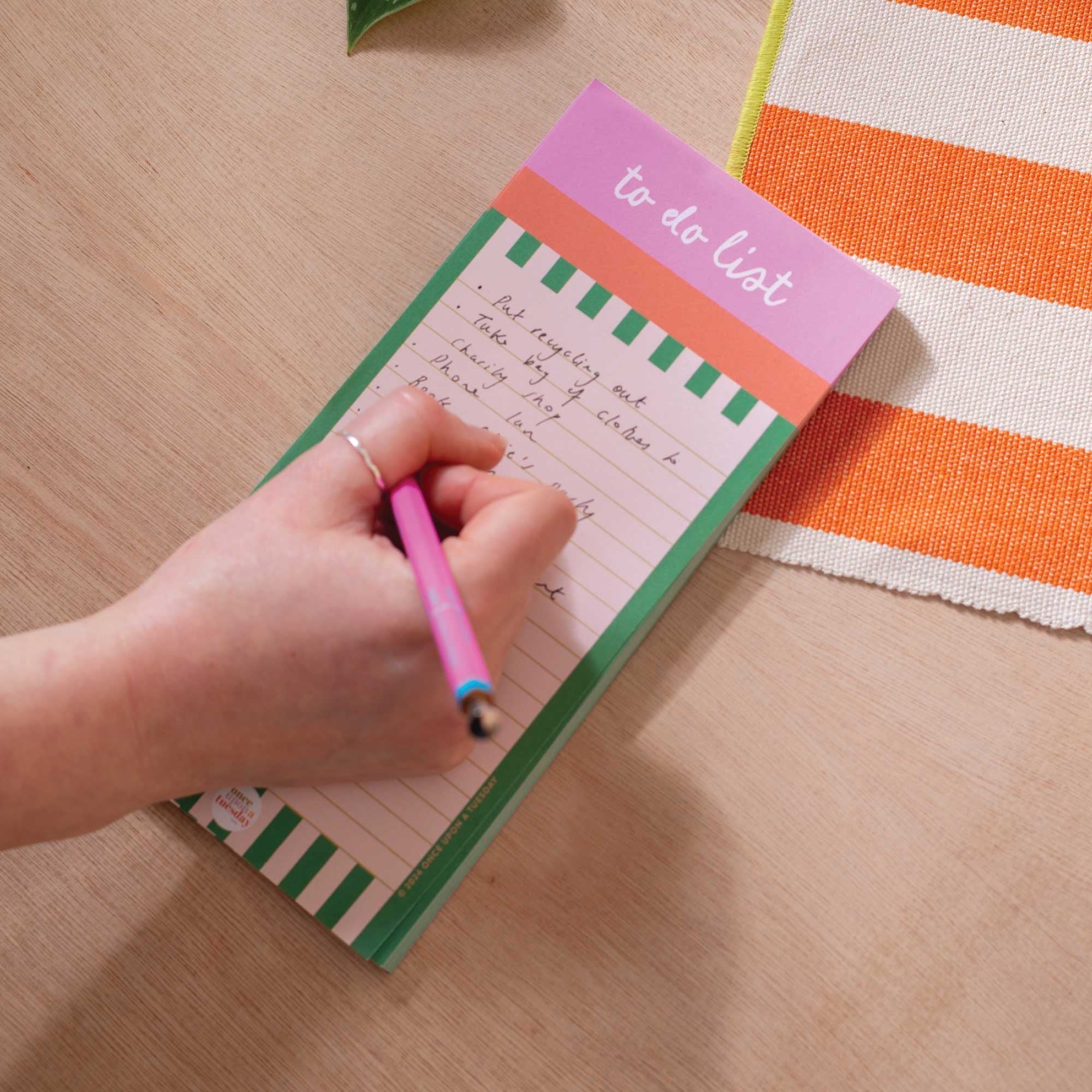Hand writing on a 'to-do list' notepad with a pink pen on a wooden surface.