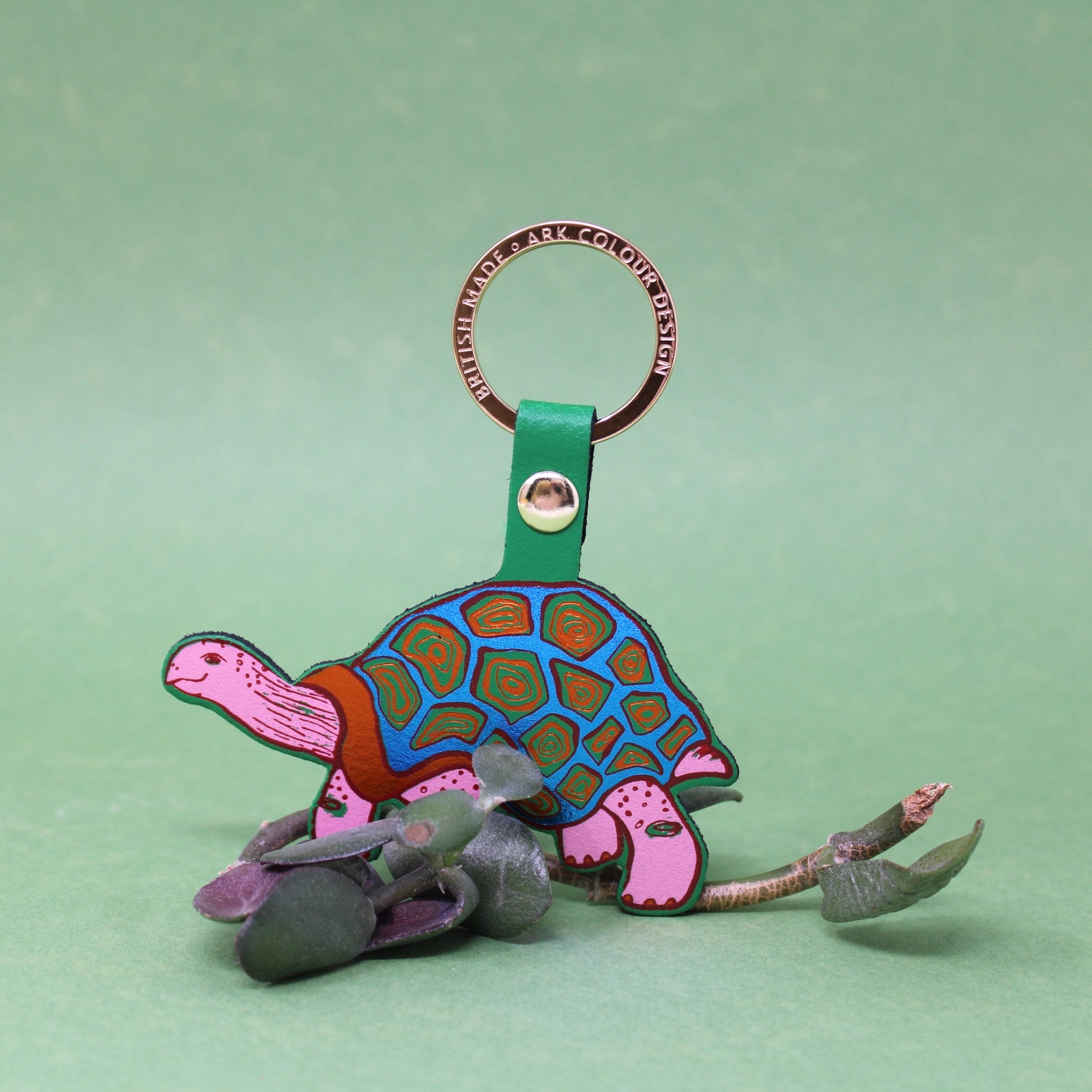Colourful tortoise-shaped keychain with a green strap on a light green background, on a branch of leaves.