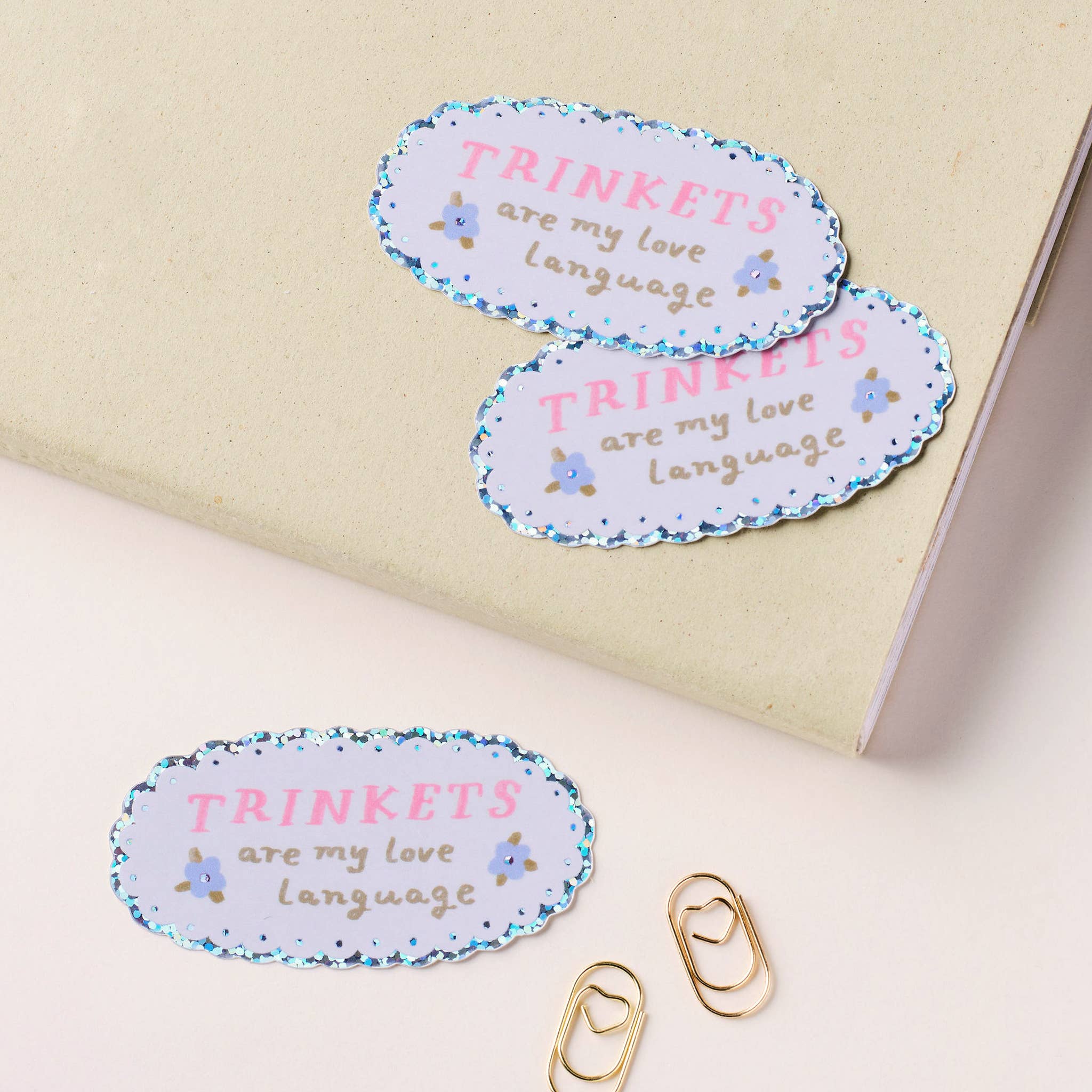 Three oval-shaped stickers with 'TRINKETS are my love language' text on a beige surface.