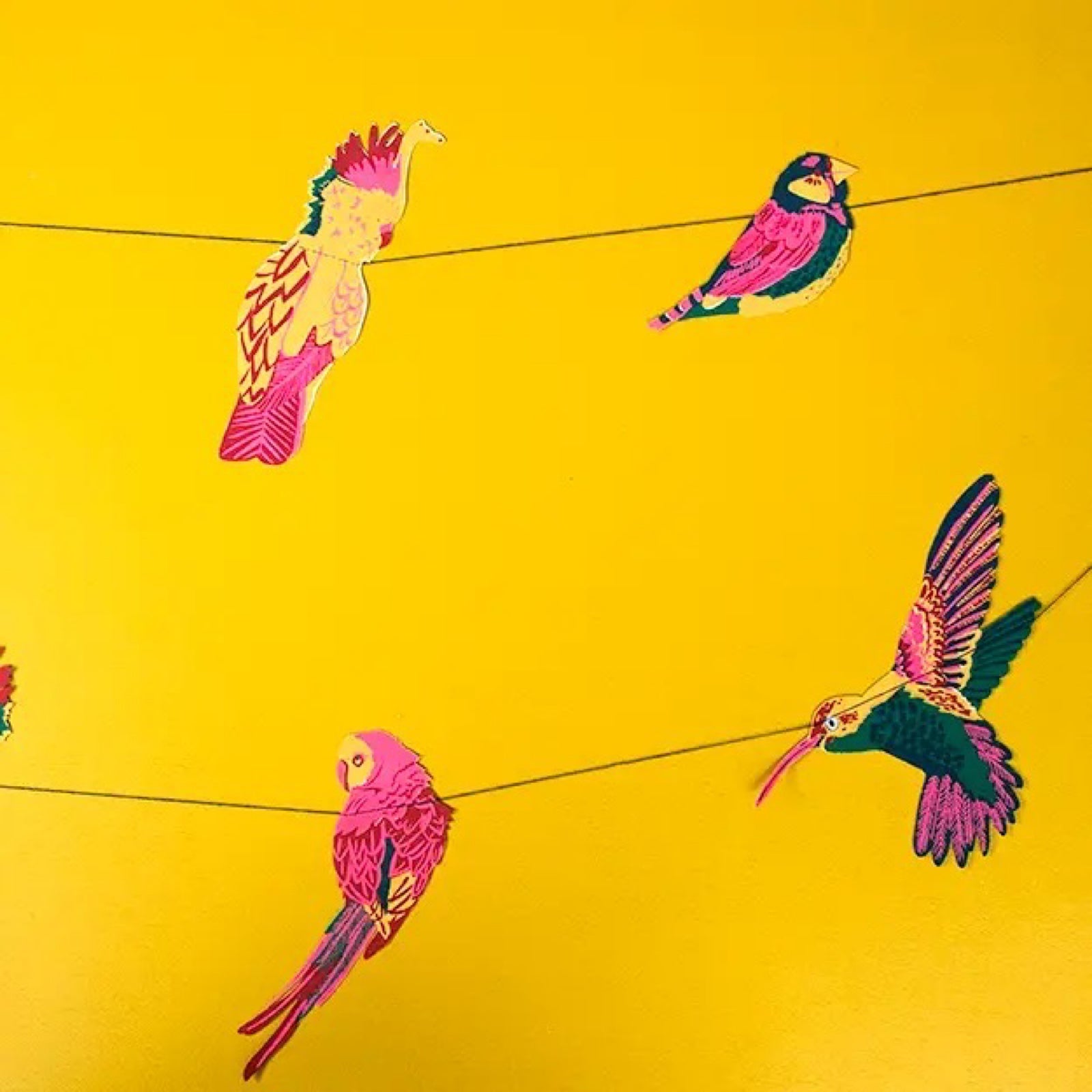 A tropical bird garland featuring ten screen-printed paper toucans, parrots and hummingbirds in bright pink, green and mustard hanging on a yellow wall.